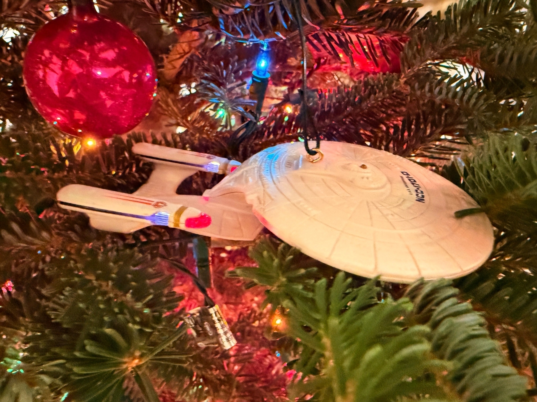 A Christmas tree decorated with colorful lights and a red ornament. Hanging among the branches is a miniature spaceship ornament resembling the Star Trek USS Enterprise.