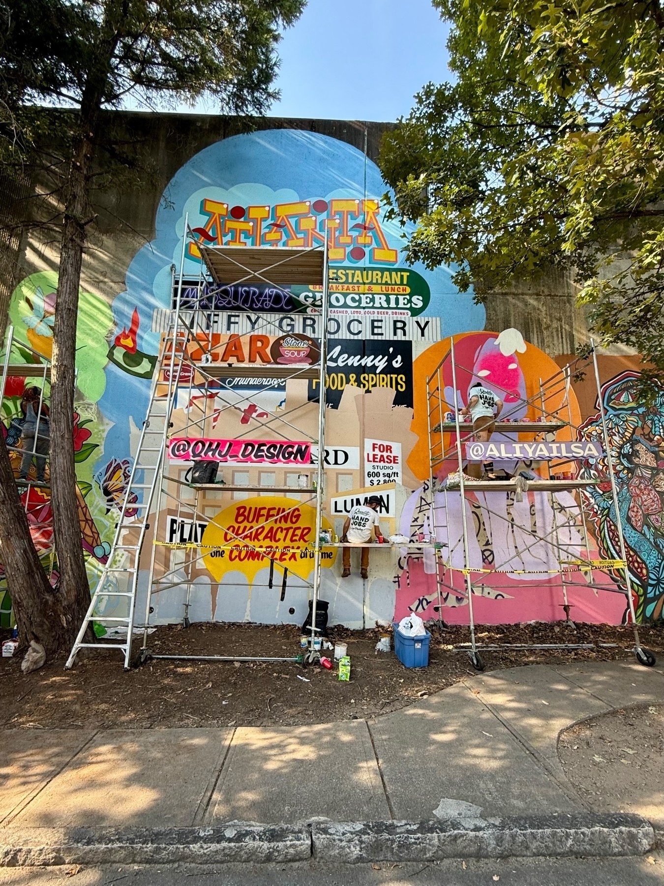 A colorful mural in progress on a wall, featuring various text and images including a restaurant sign, a grocery advertisement, and artistic designs. Two ladders and scaffolding are set up, and some painting supplies and materials are scattered on the ground.