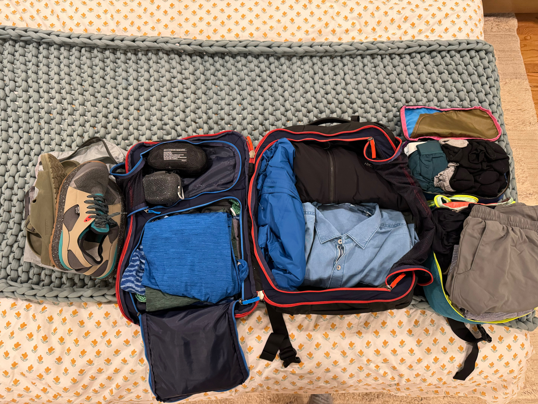 An open suitcase on a bed with neatly organized clothes and a pair of shoes beside it. The suitcase contains various clothing items including shirts and pants, and utilizes packing cubes for organization.