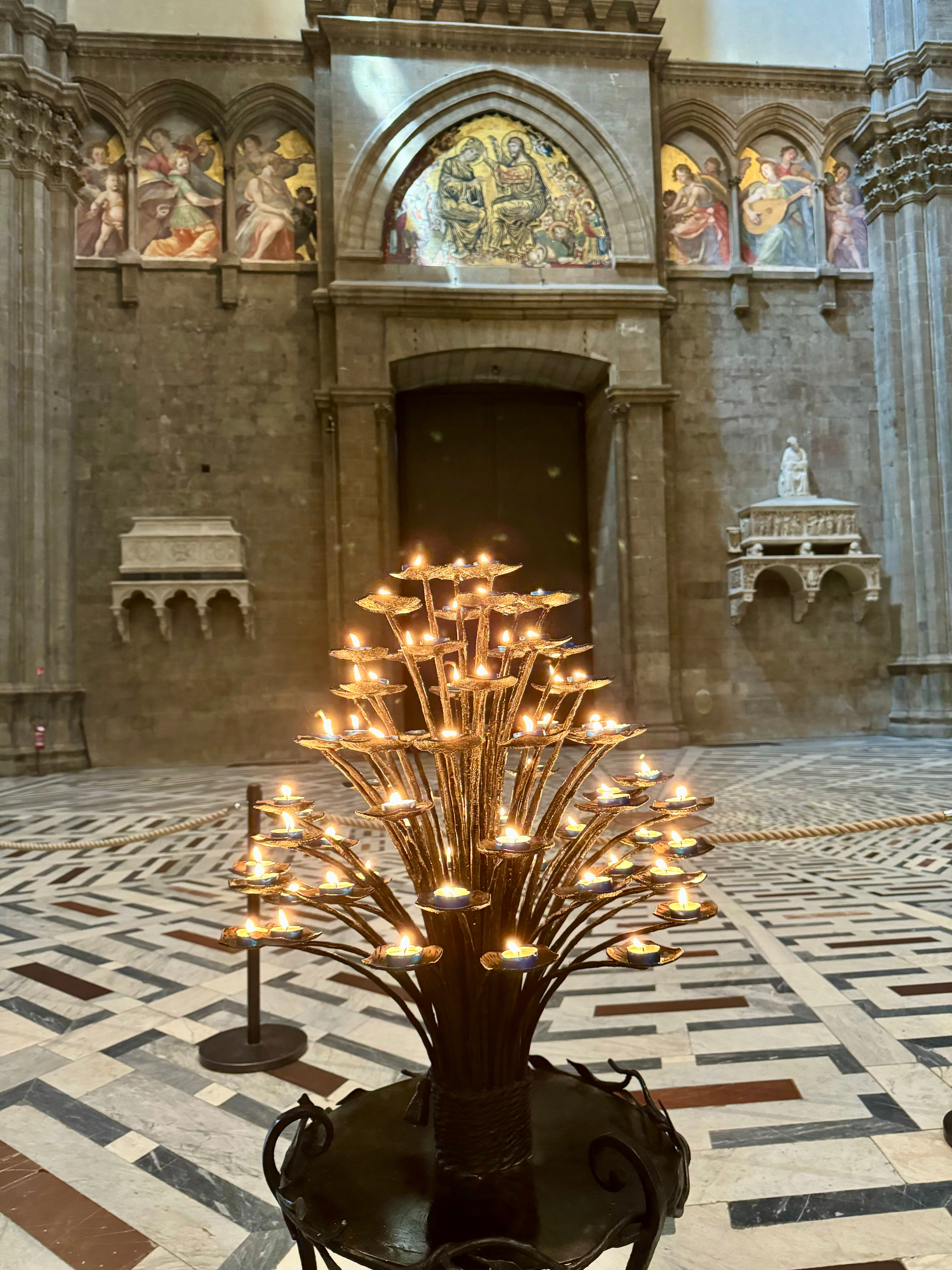 An ornate candle stand with multiple lit candles in a cathedral. The background features a large, arched doorway with a mosaic of religious figures above it, flanked by stone columns and additional wall decorations, including painted frescos and statues. 