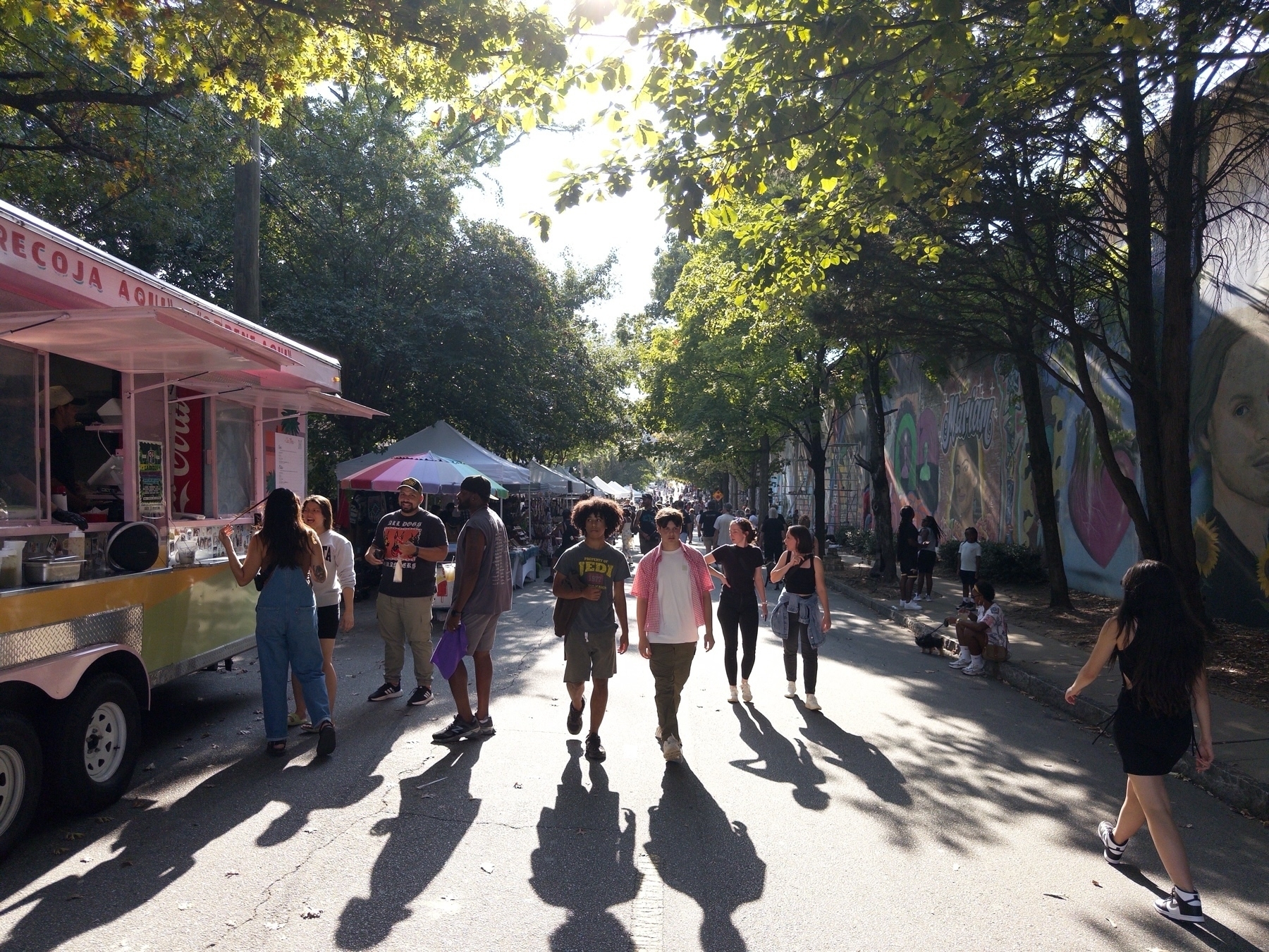 A bustling street scene with people walking along a road lined with trees and street art. A pink food truck is parked on the left with several people ordering. The atmosphere is lively and the sun is casting long shadows on the ground.