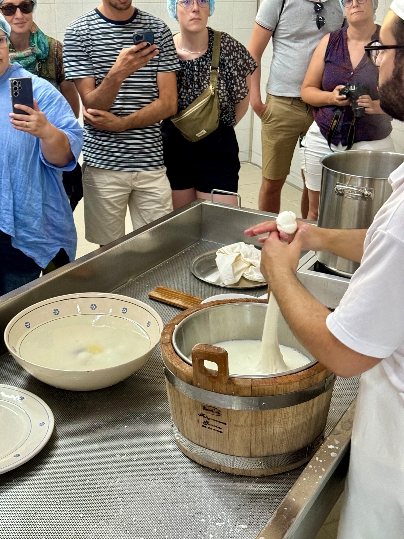 A group of people watching a demonstration of traditional cheese-making. The instructor is handling fresh cheese curds, pulling them into a stretchy mass. Various bowls and a wooden bucket with a milk-based liquid are on a metal countertop. 