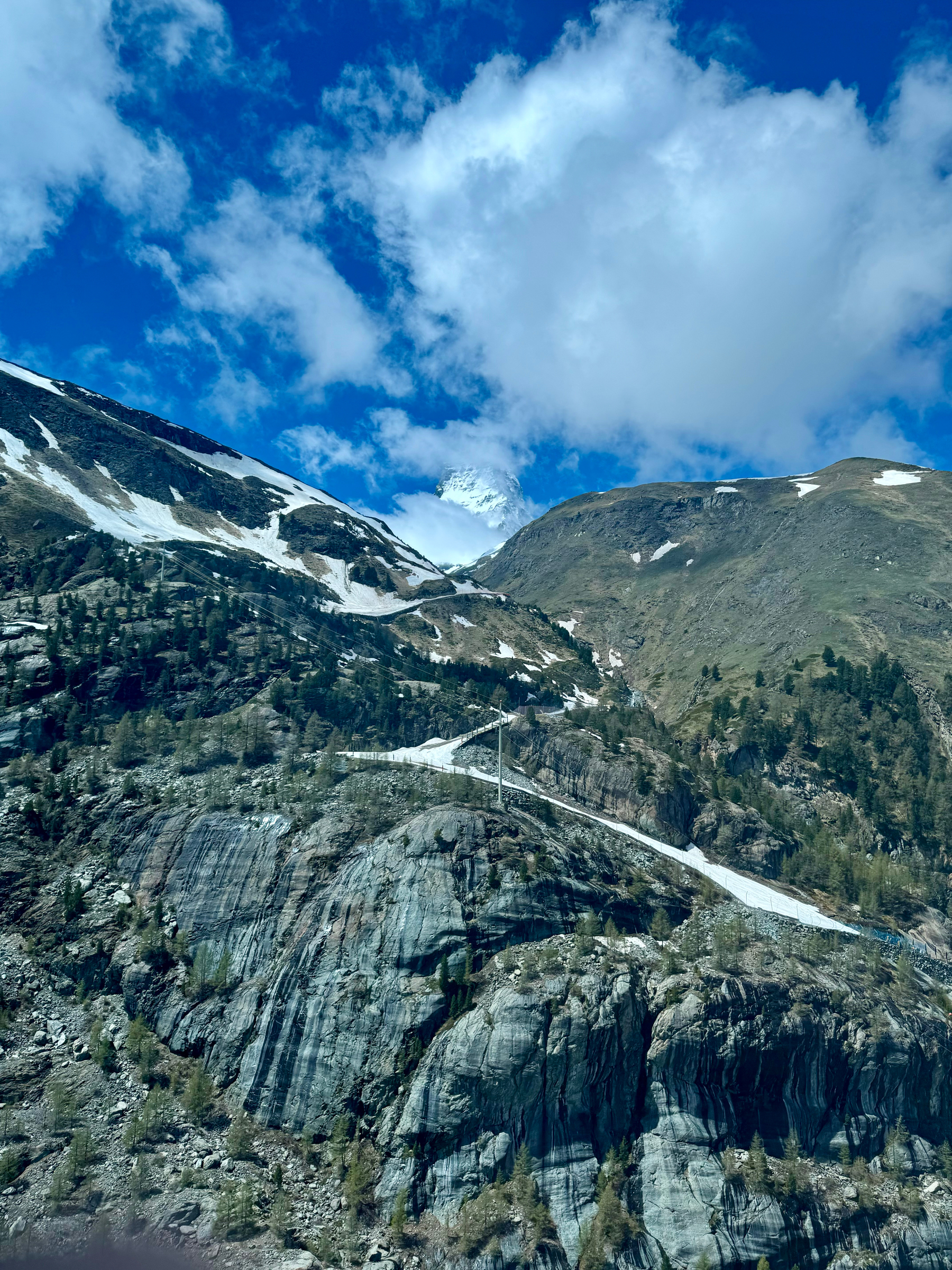 Mountainous landscape with a winding road, snow patches, rocky terrain, and a blue sky with clouds.