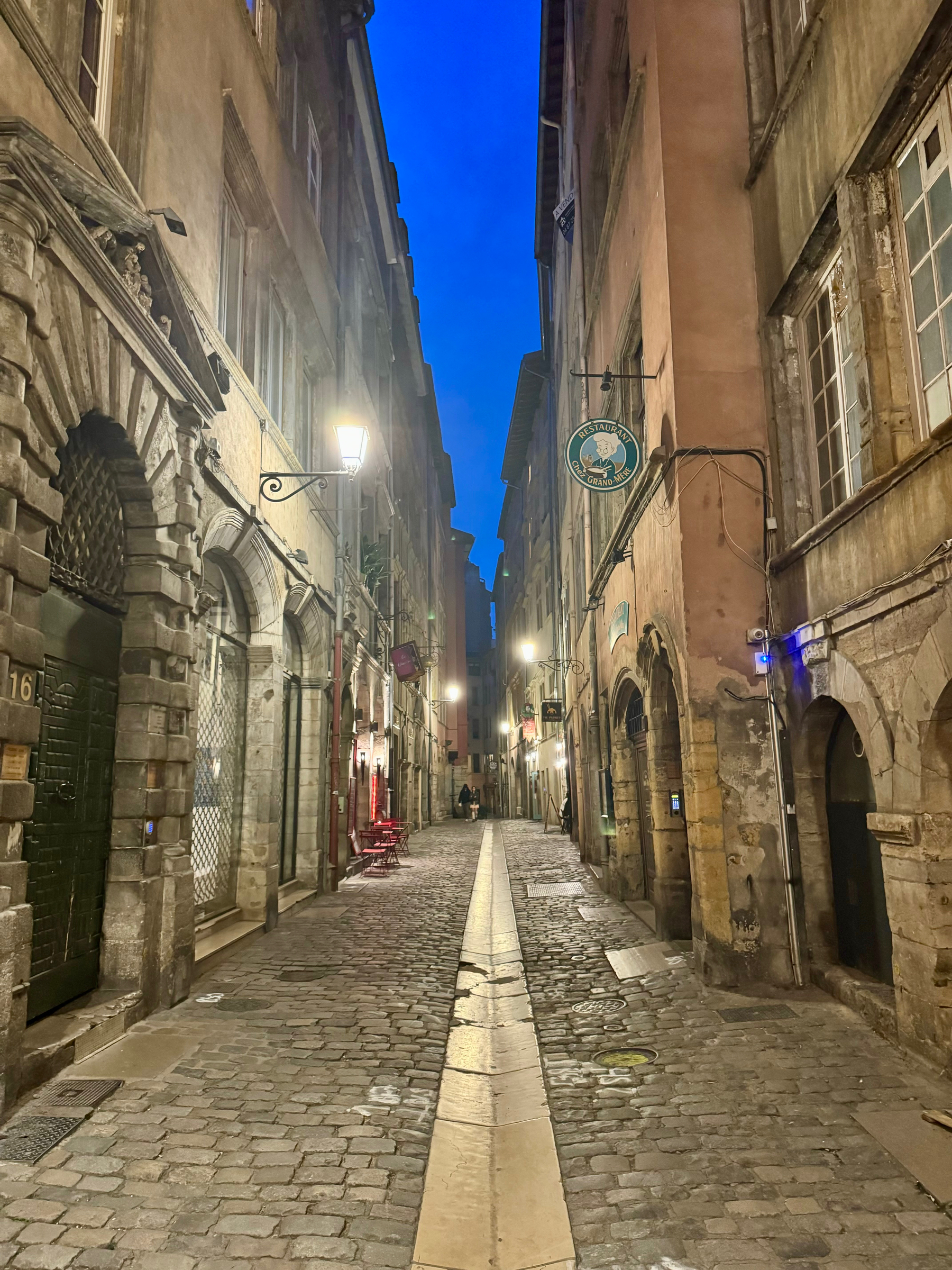 A narrow cobblestone street lined with old buildings and illuminated by street lamps at twilight, signs for businesses hang on walls, and a few people are visible in the distance.