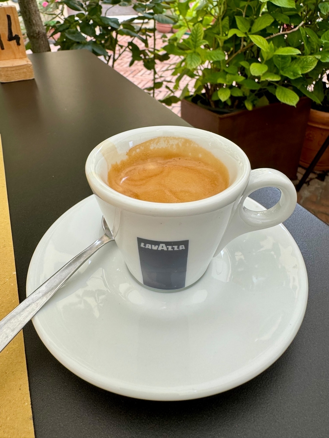 A Lavazza-branded white espresso cup and saucer filled with espresso, placed on a table. A metal spoon rests on the saucer. In the background, there are green plants and a wooden table number stand with the number 14.