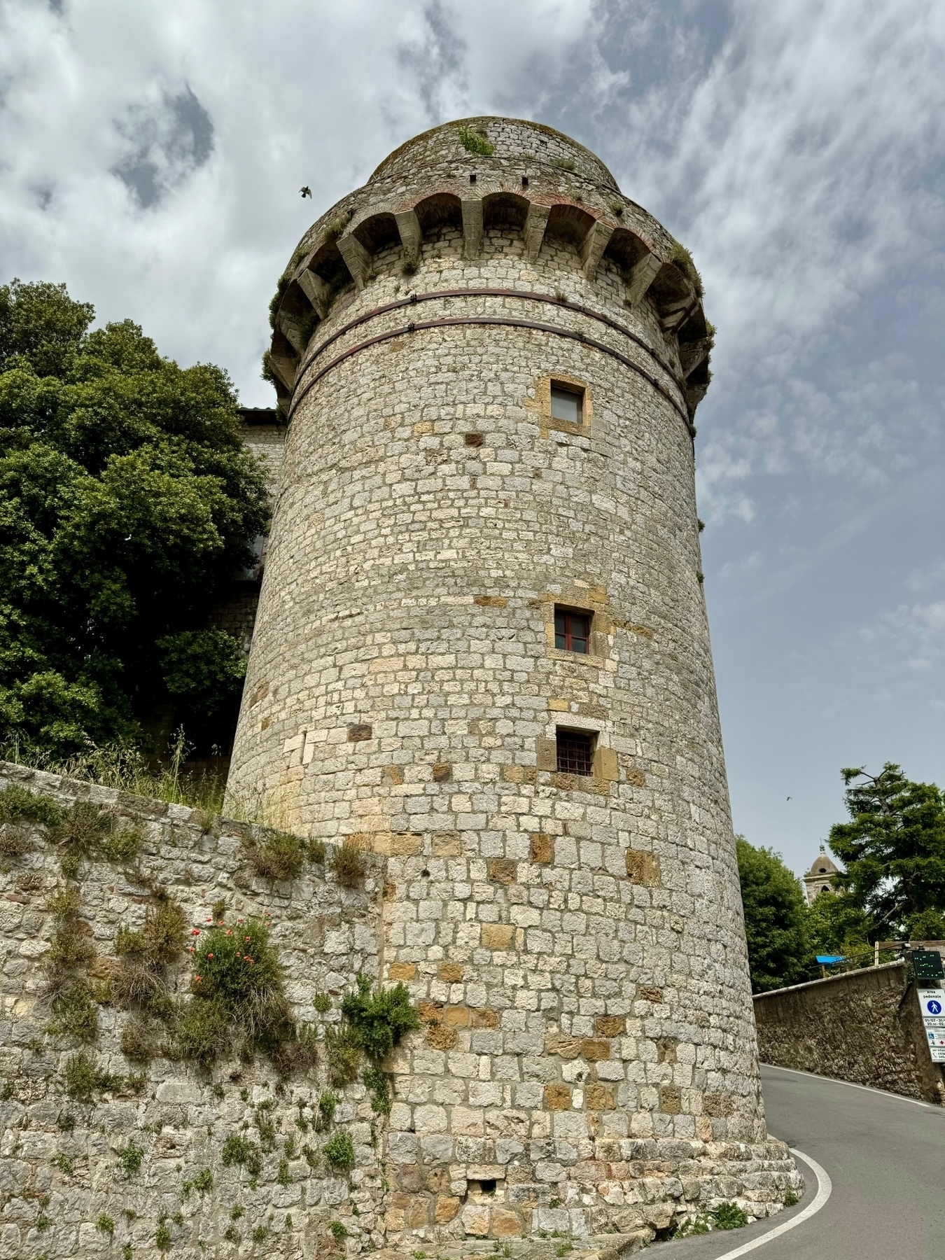 A medieval stone tower with a cylindrical shape, featuring small windows, a conical roof, and an overhanging battlement. The tower is situated next to a winding road with trees and vegetation nearby. A partly cloudy sky is visible in the background. 