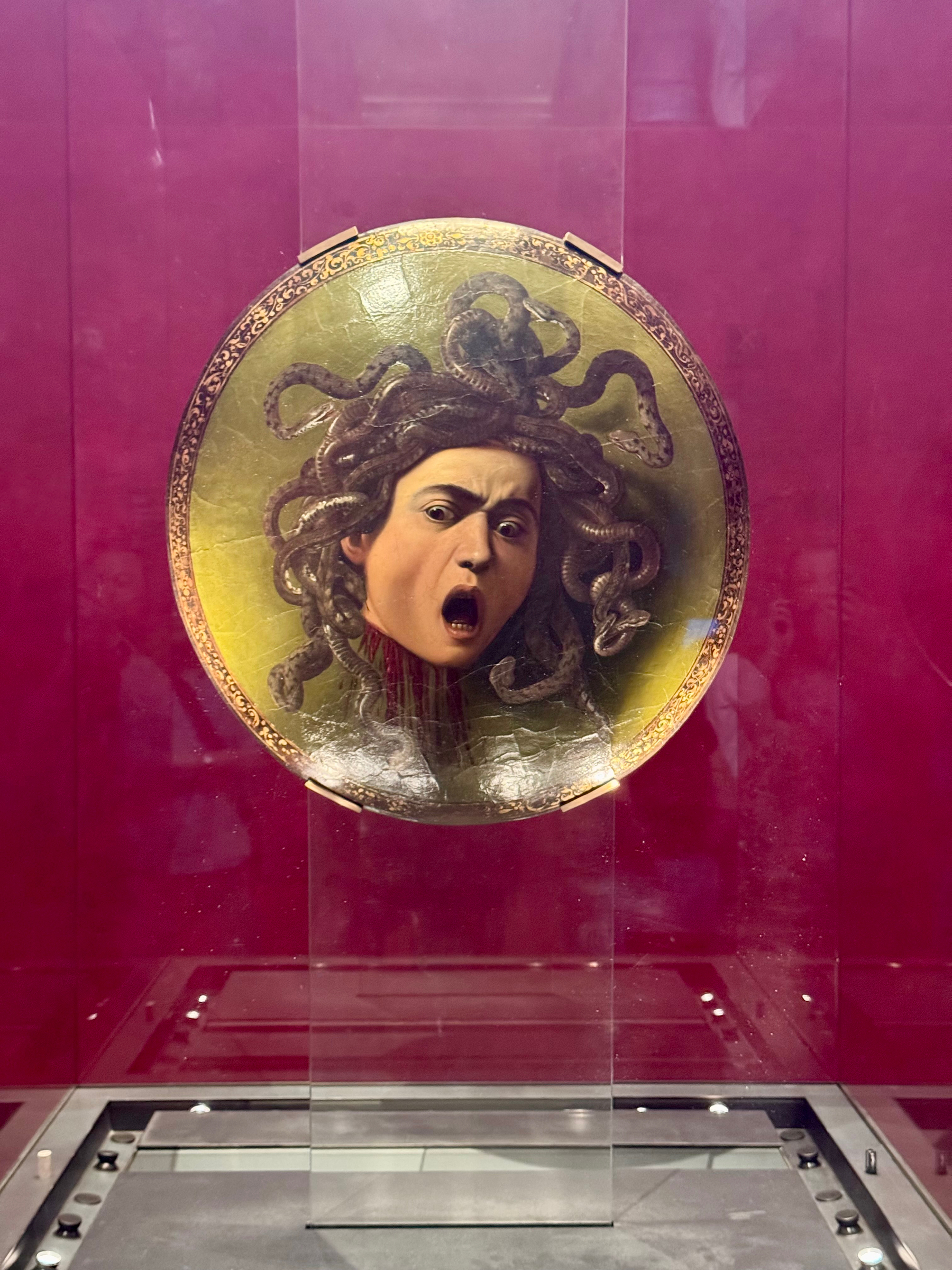 A painting depicting the head of Medusa, with writhing snakes for hair and a pained expression on her face, displayed in a glass case against a red background. The artwork is mounted on a round shield-like surface.