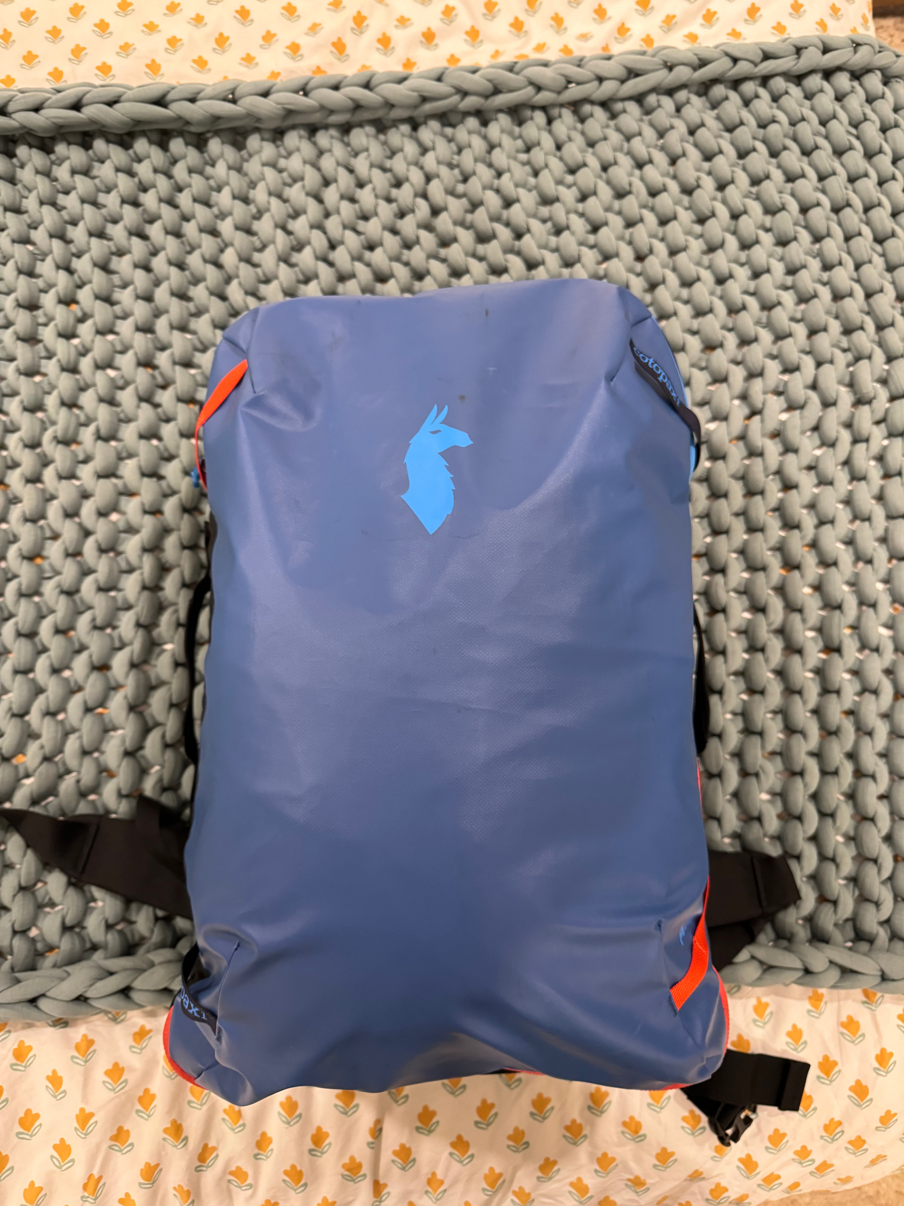 A blue travel backpack with a silhouette of an alpaca on it, placed on a gray knitted blanket over a patterned sheet.