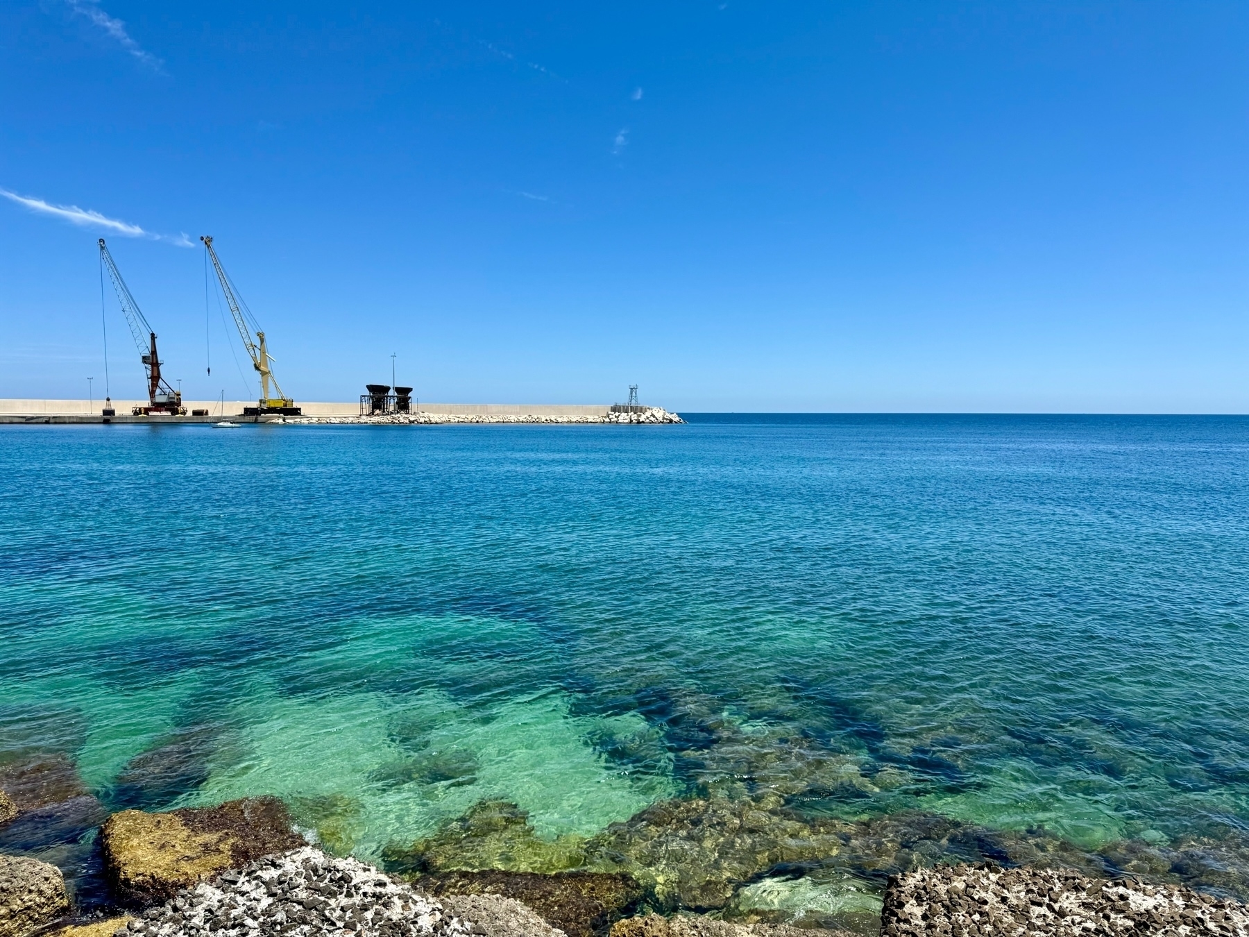 A scenic view of a harbor with crystal-clear turquoise water and a rocky shoreline. There are two cranes on the pier, extending into the blue sky, which is clear with only a few wispy clouds. The horizon line separates the sea from the sky. 