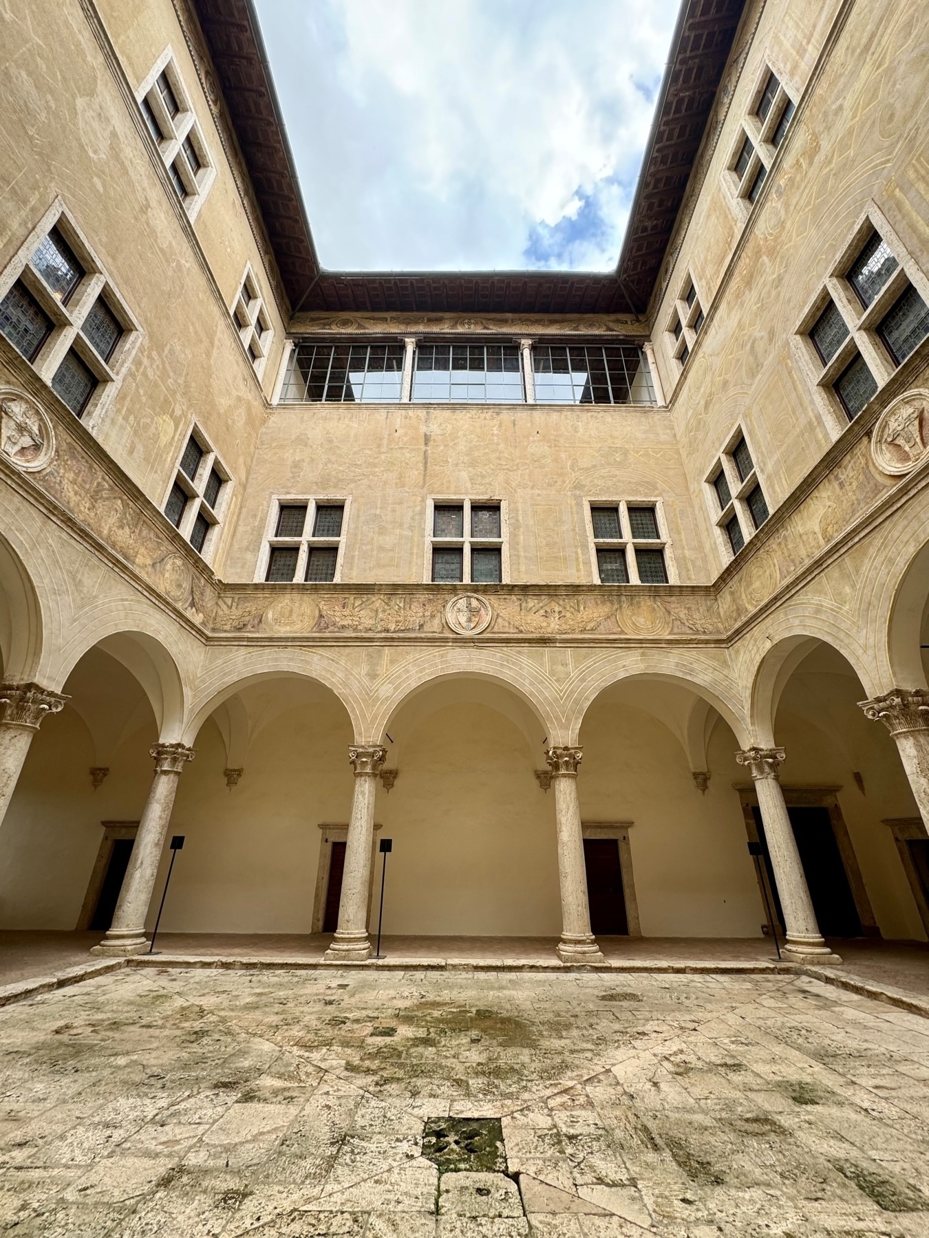 The image depicts the interior courtyard of a historic building, featuring an open atrium with arched columns and two levels of windows. The courtyard has a stone floor with weathered and cracked tiles. The upper level has decorated walls and windows. 