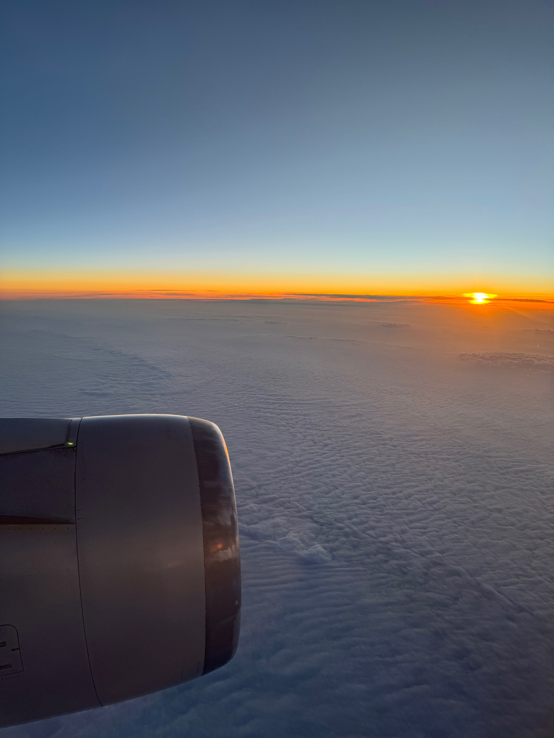 Airplane wing and engine against a backdrop of a sunrise sky with clouds below.