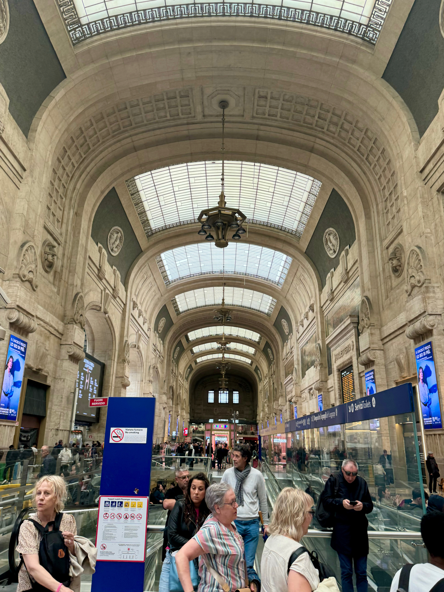 Interior of a grand train station with high arched ceilings, ornate architectural details, and large windows providing natural light. Several people are walking, standing, and using escalators in the busy concourse. Signs with information and advertisements are visible throughout the scene. 
