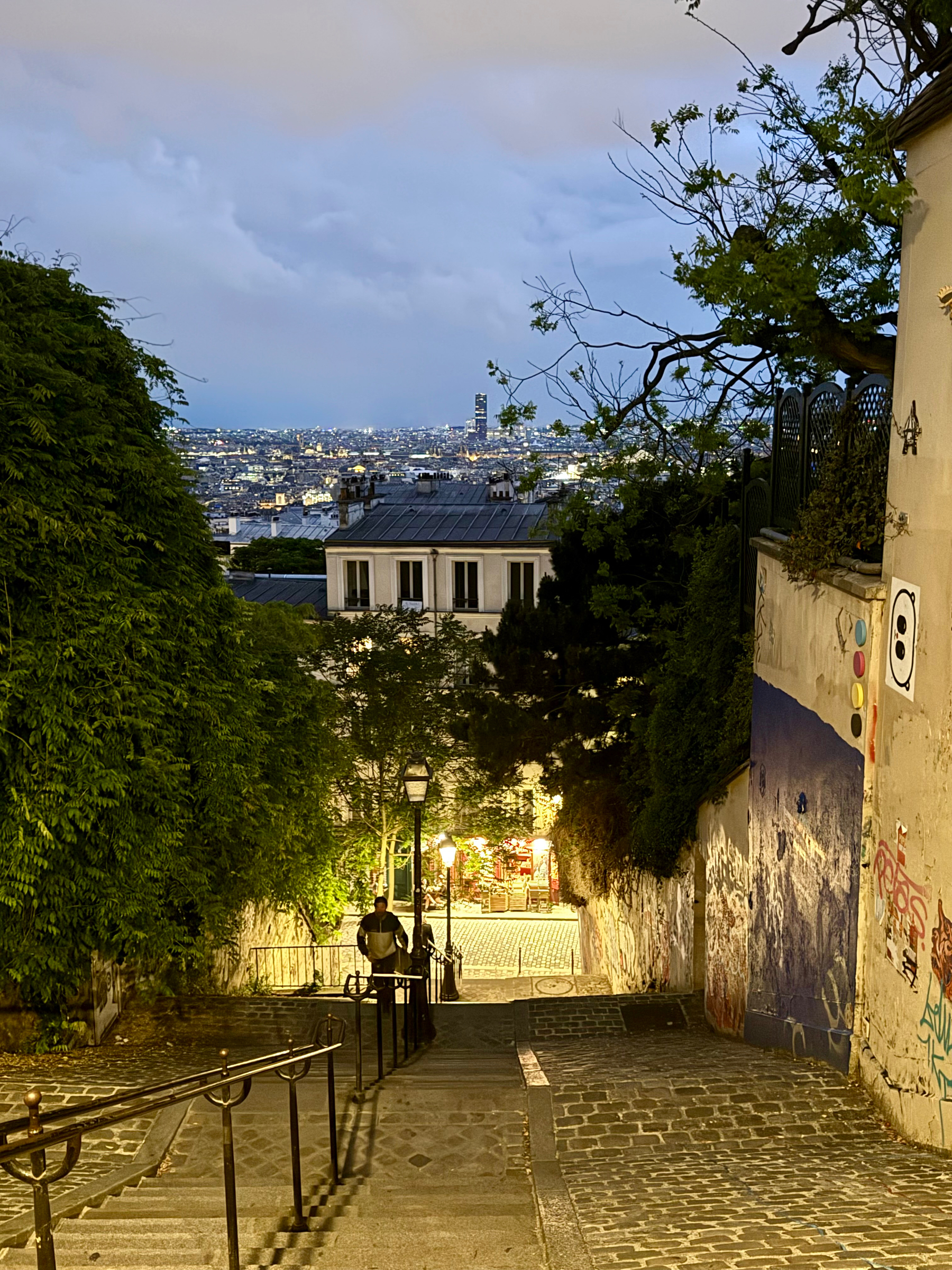 A nighttime view of a cobblestone staircase descending towards a brightly lit street, with city lights in the background, flanked by trees and walls with graffiti, and a person walking up the stairs.