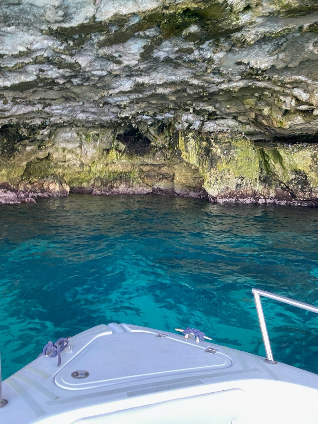 A boat is partially visible as it enters a rocky cave with a low ceiling. The water in the cave is a clear, vibrant blue and the cave walls are covered in green moss and algae.