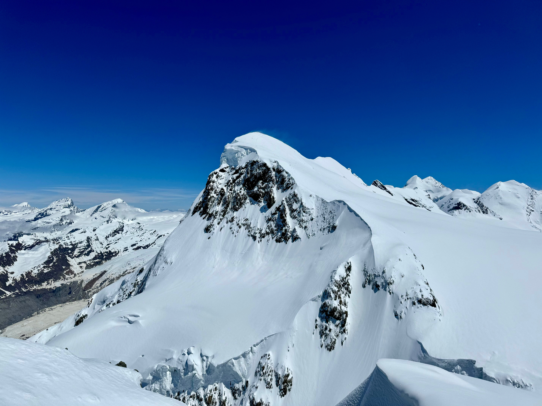 A snow-covered mountain peak under a clear blue sky with surrounding alpine scenery.