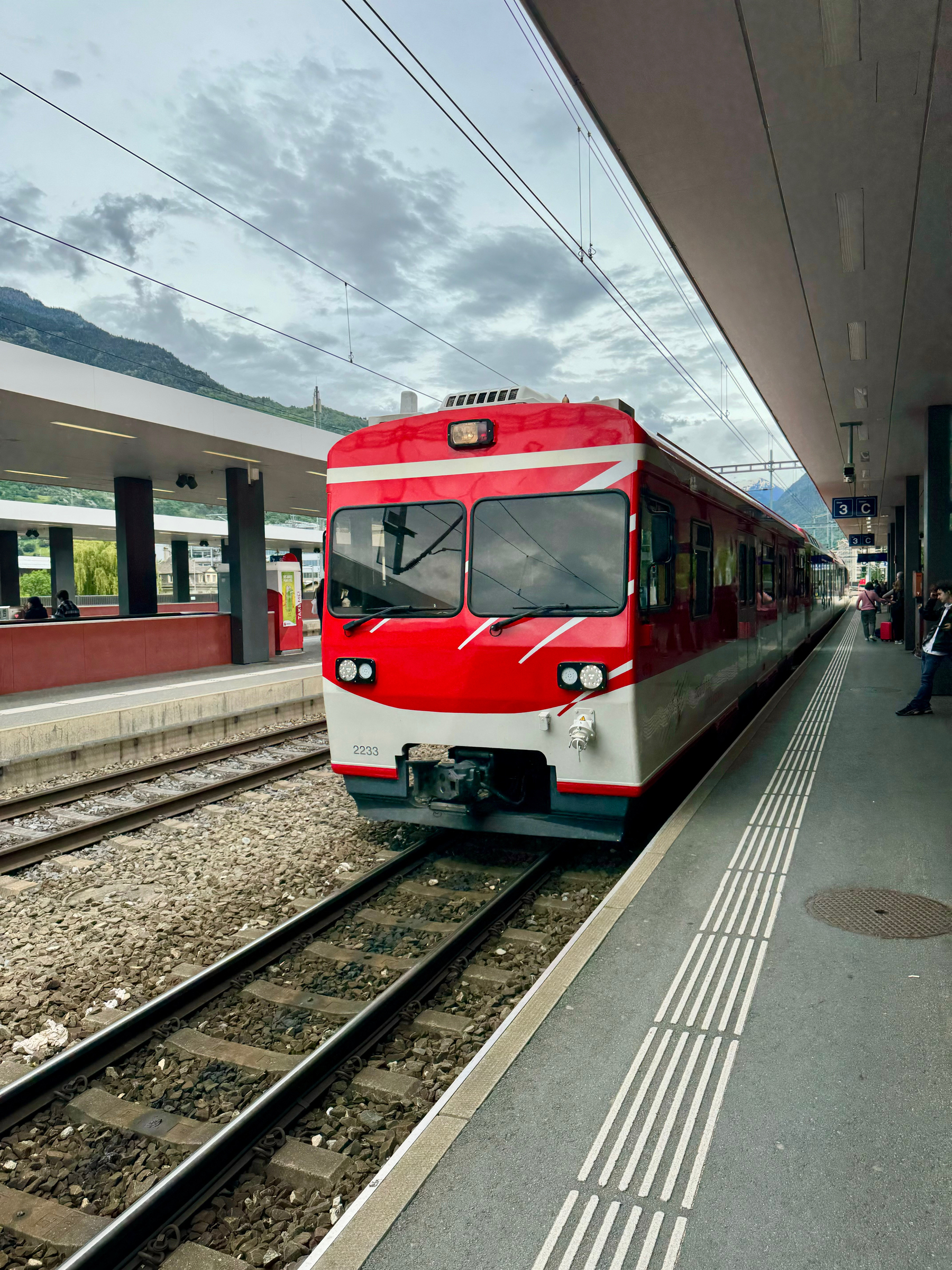 A red and white train at a platform with mountains in the background and a partly cloudy sky above.
