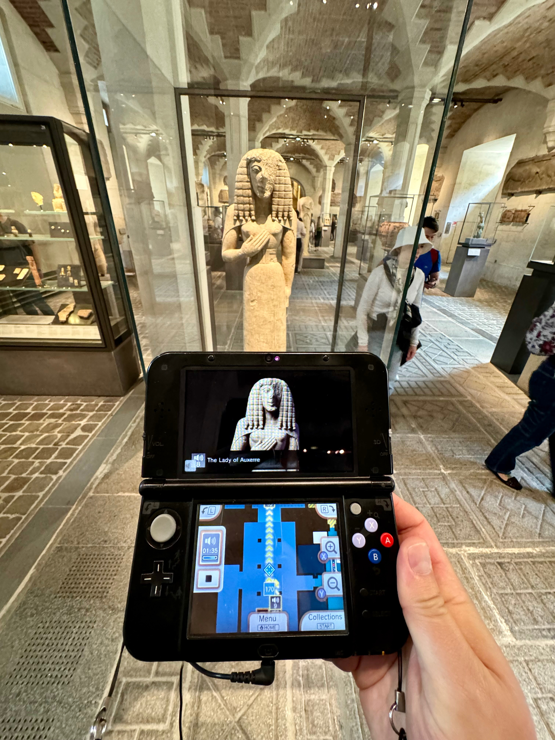 A handheld Nintendo 3DS gaming system is displaying a digital image of an ancient statue titled “The Lady of Auxerre,” perfectly lined up with the actual statue visible in a glass display case in the background of a museum. 