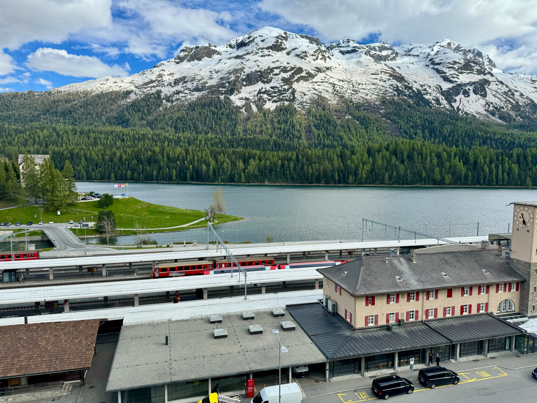 A train station with red trains, a lake, a forest, and snow-capped mountains in the background.