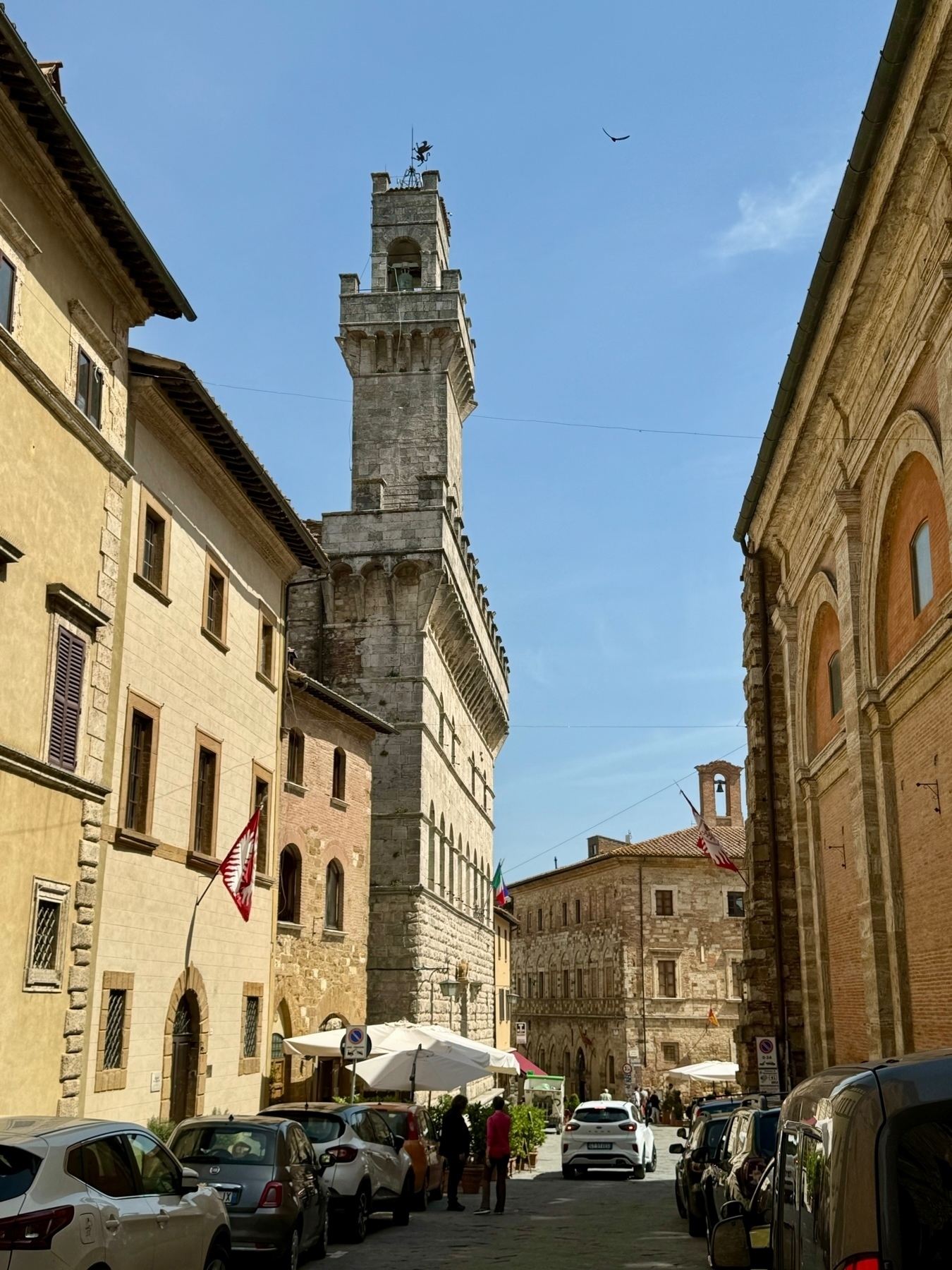 View of a narrow street in an old town, flanked by historic buildings with stone facades. In the center is a tall clock tower with a bell at the top. A few people are walking or sitting at an outdoor café with white umbrellas.