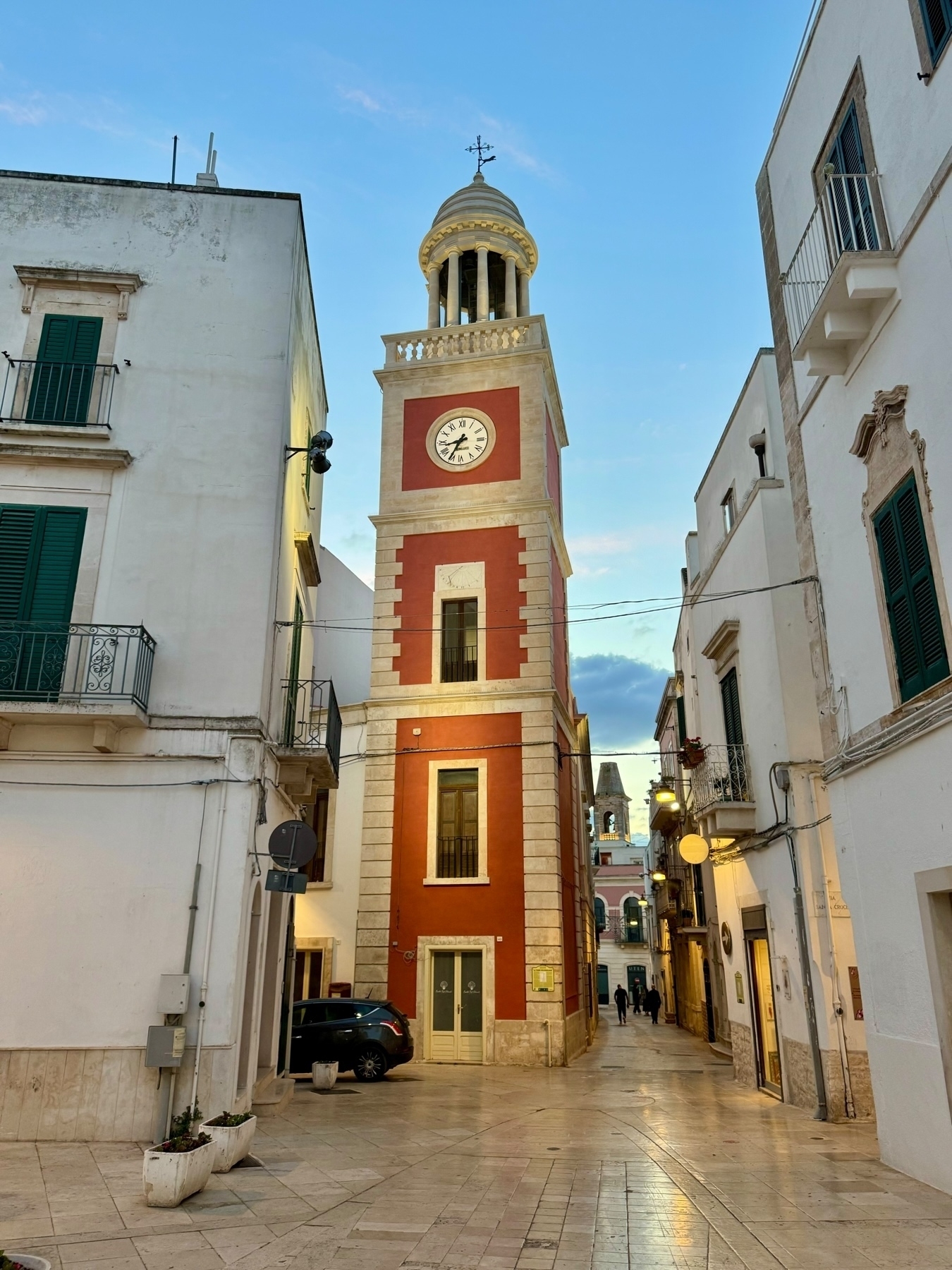 This image features a street view of a picturesque European town during dusk. The focal point is a tall, red clock tower with a white clock face and a dome roof. The tower is flanked by whitewashed buildings with green shutters and balconies. 