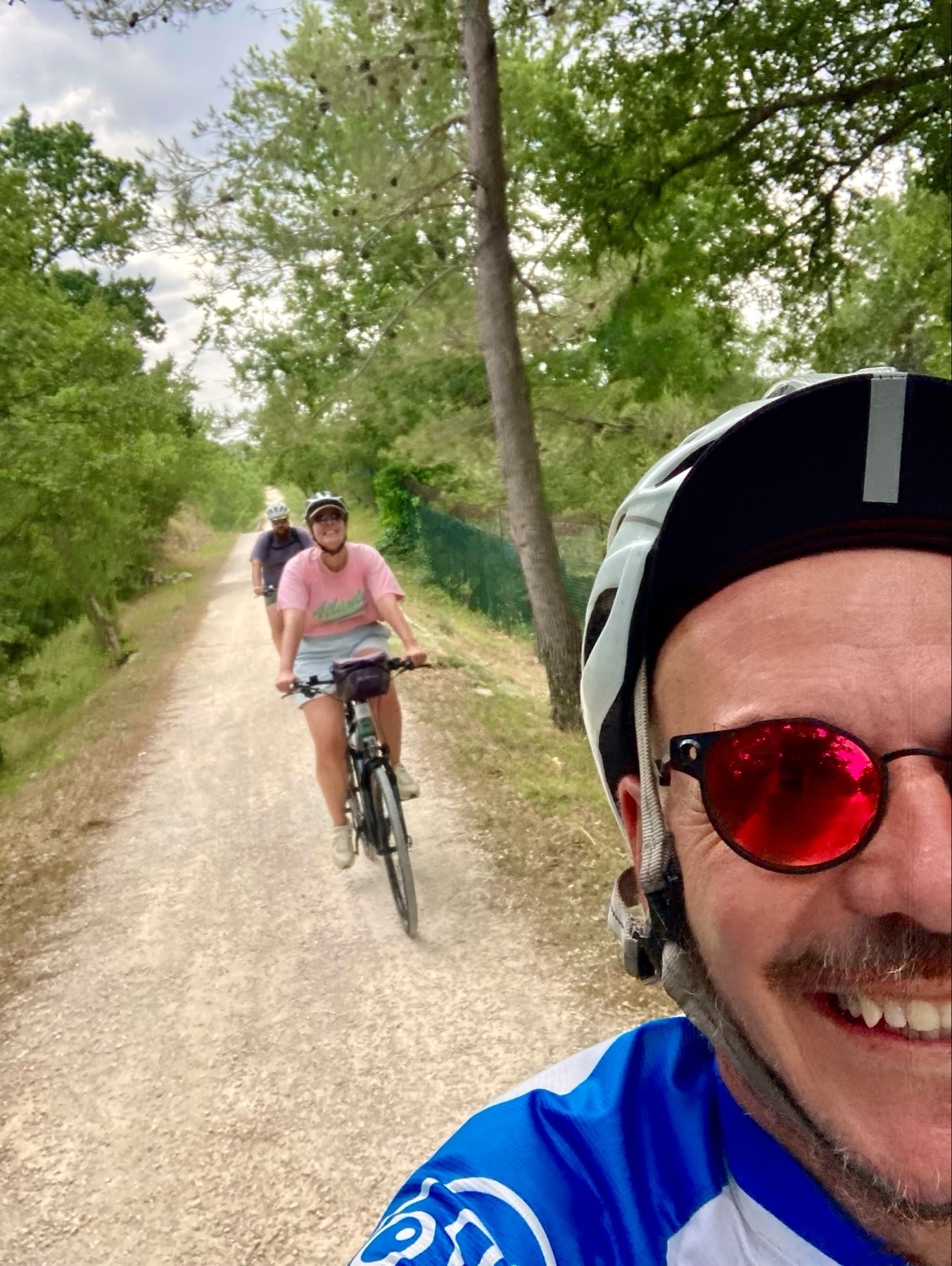 A group of three cyclists riding on a dirt trail surrounded by trees and greenery. The foreground features a smiling cyclist in a blue and white jersey with red-tinted sunglasses, taking a selfie. Two other cyclists can be seen in the background wearing helmets.