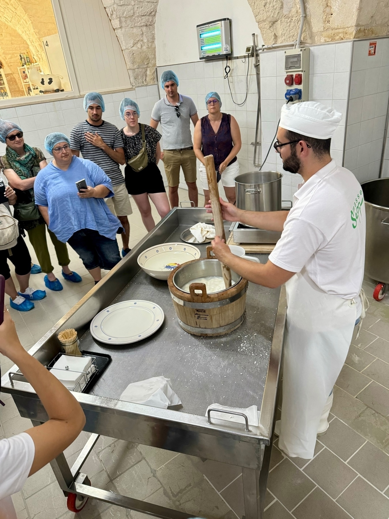 A group of people wearing hairnets and protective shoe covers are observing a cheesemaker demonstrate cheese-making. The cheesemaker, wearing a white uniform and hat, is stirring curds in a wooden bucket on a metal table.