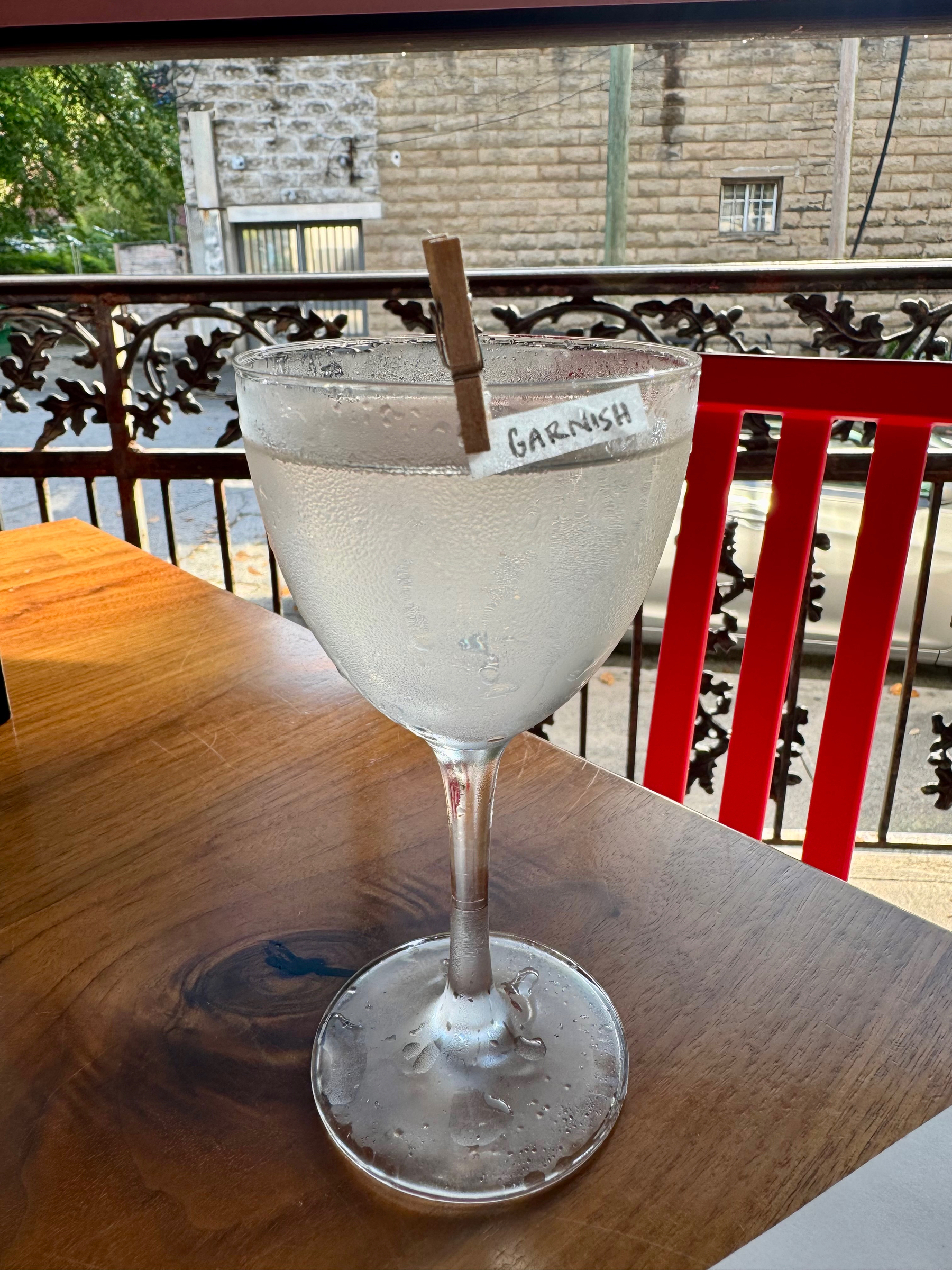 A chilled beverage in a stemmed glass on a wooden table with the word “GARNISH” on a clip attached to the glass. In the background, there’s a balcony with a decorative railing overlooking a street and a stone building.