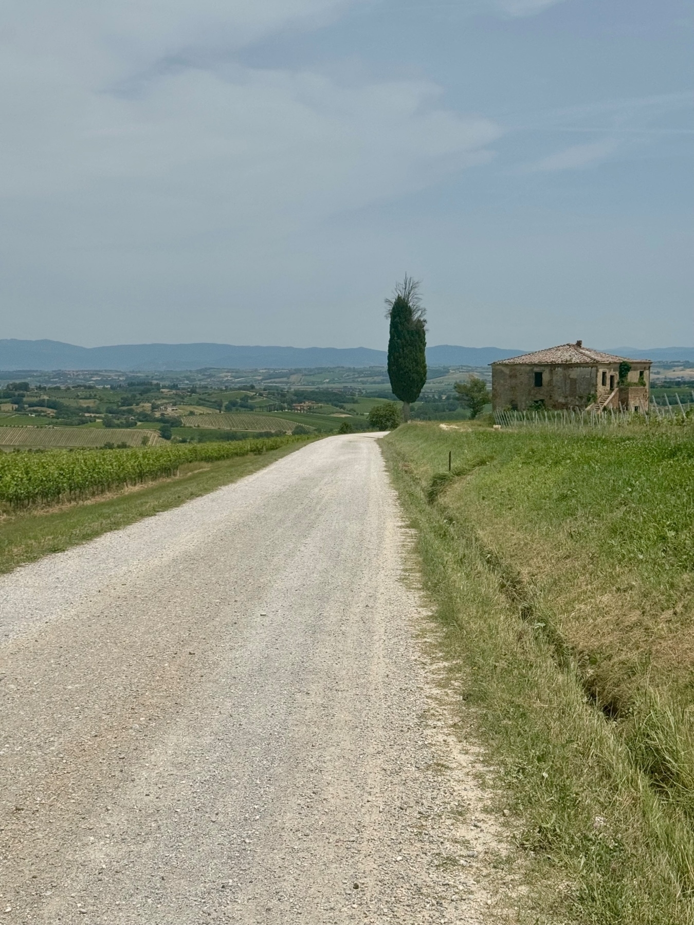 A gravel road running through a rural landscape with greenery and rolling hills in the background. On the right side of the road, there is an old, rustic stone building with a tiled roof, and a tall, narrow cypress tree stands nearby. 