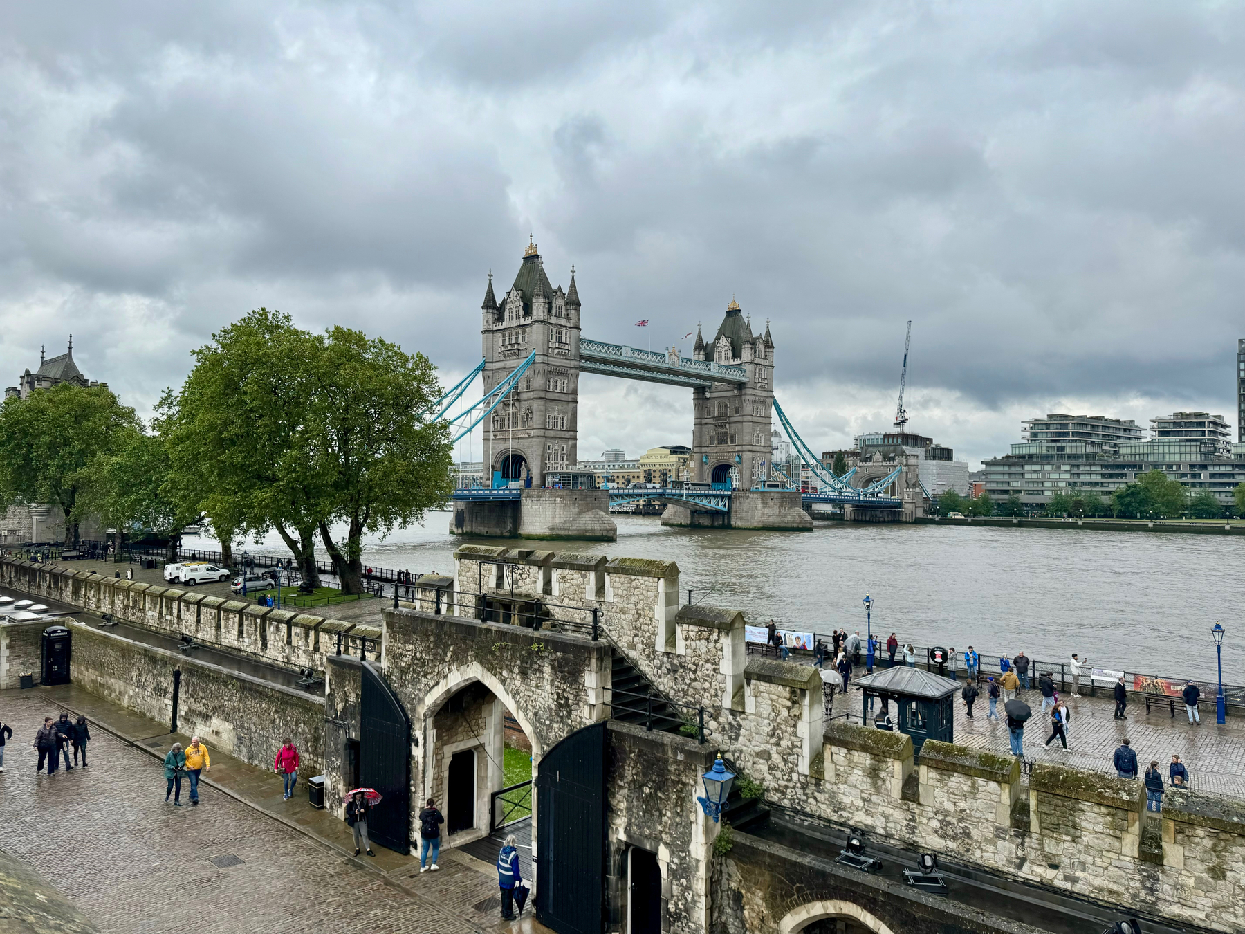 Tower Bridge over the River Thames in London, viewed from a high vantage point showing the bridge’s twin Gothic towers, with people walking along the riverbank and part of the historical Tower of London visible in the foreground.