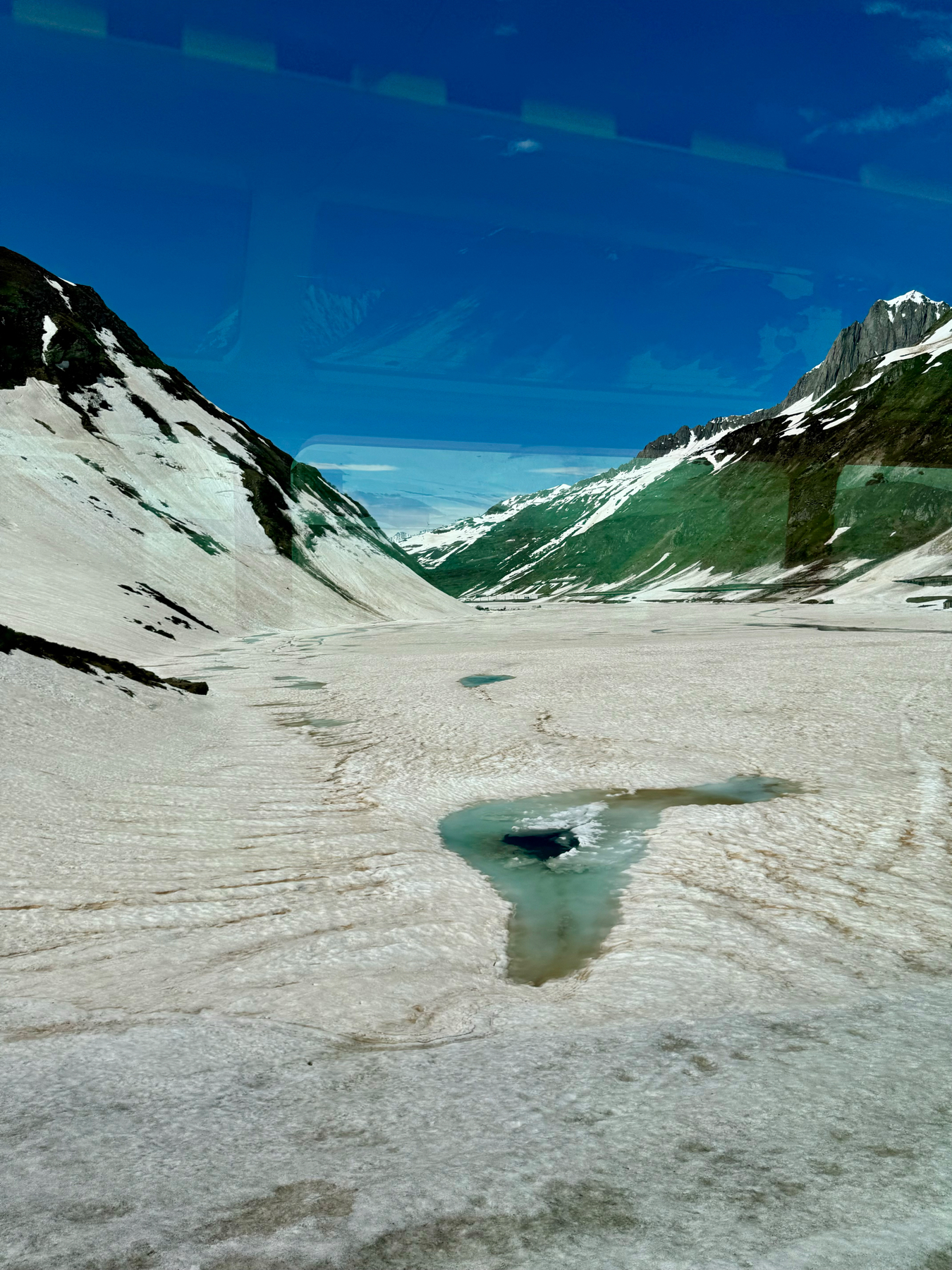 A glacier with a snow-covered surface featuring meltwater pools against a backdrop of mountain slopes and a clear blue sky.