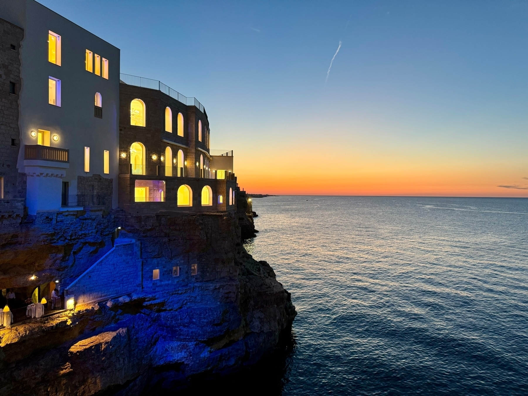 Coastal building with illuminated windows perched on a cliffside at sunset, overlooking a calm sea with a pastel-colored sky in the background. The lower part of the building and cliff are softly lit in blue.