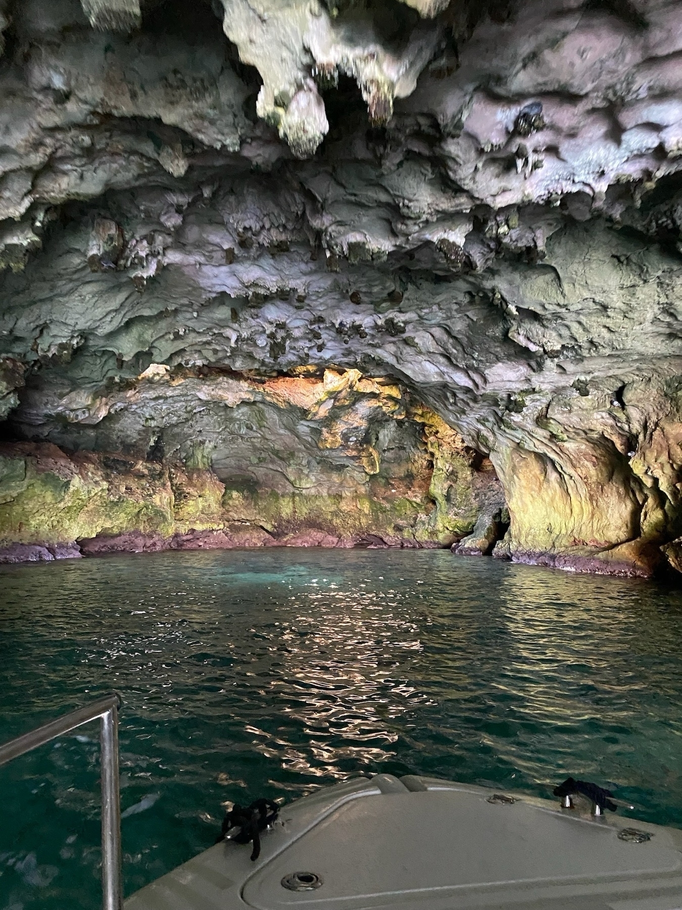 A photo taken from a boat inside a cave with greenish-blue water. The cave walls and ceiling are rocky, with varying shades of green, purple, and brown due to mineral deposits and lighting.