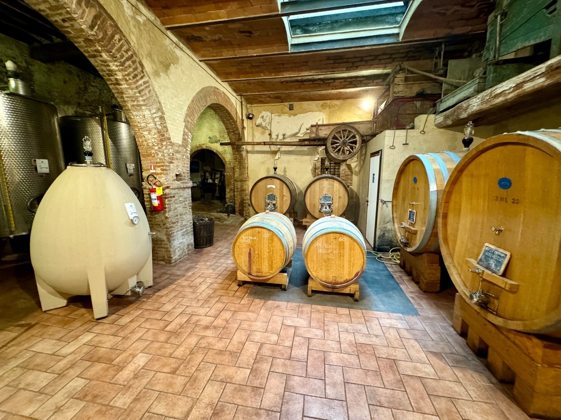This image shows the interior of a winery cellar with various barrels and tanks used for storing and aging wine. There are large wooden barrels, a ceramic fermentation tank, and stainless steel vats. The space features brick arches, rustic decorations, and industrial elements. 