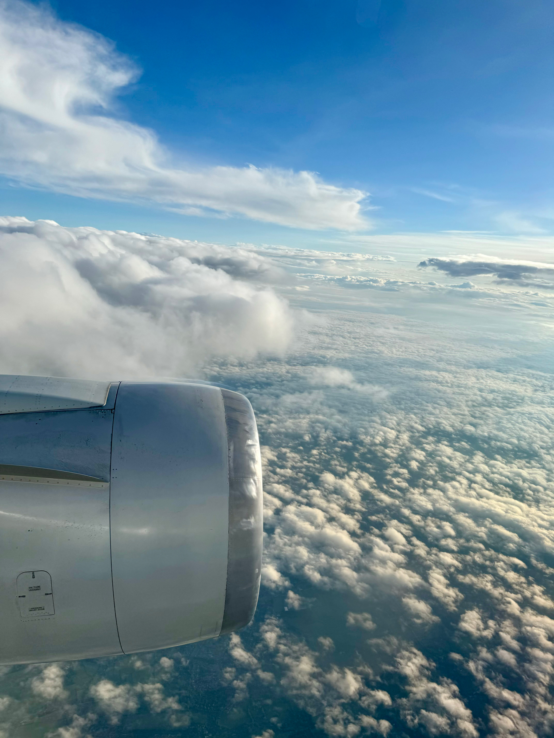 View from an airplane window showing the engine and wing over a seascape of clouds under a blue sky.