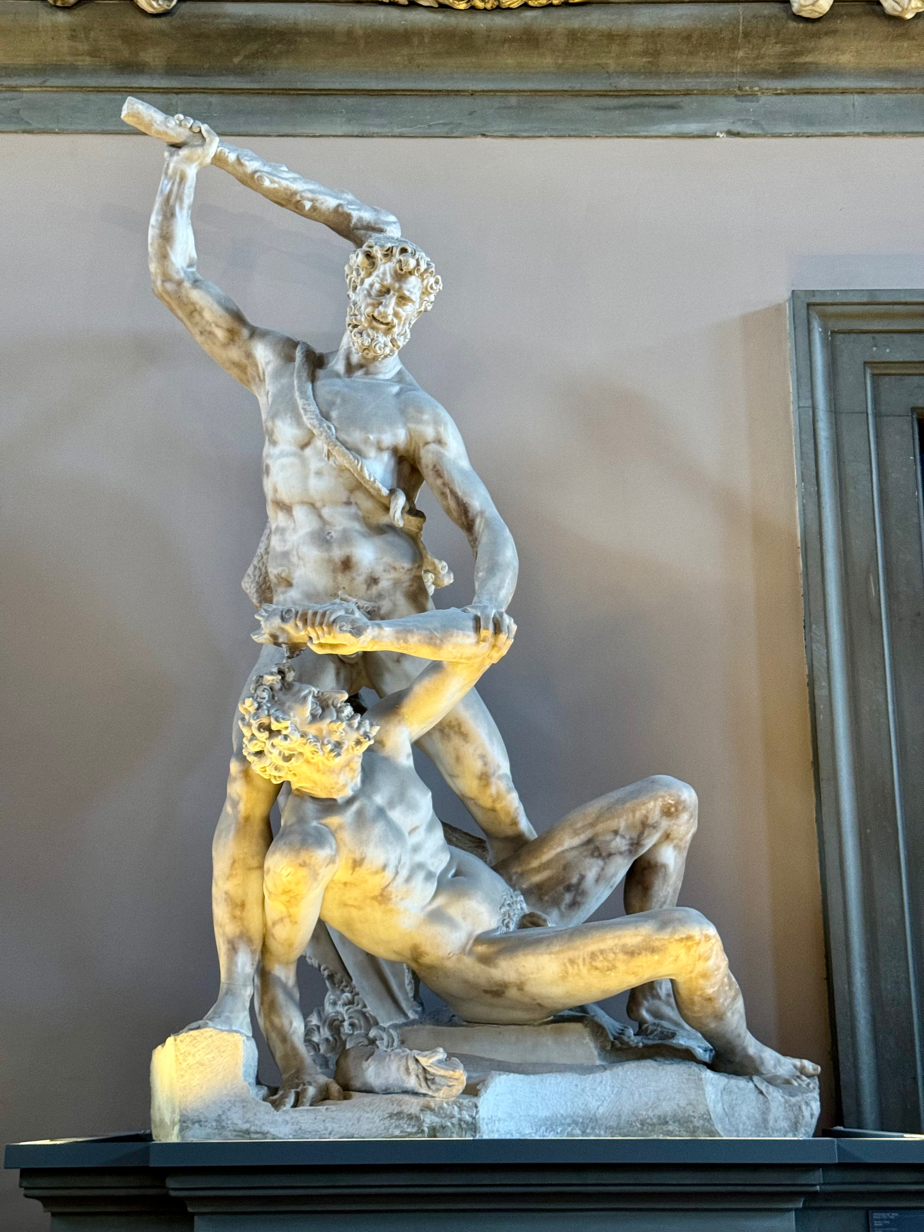 A marble sculpture depicting two male figures in a dynamic struggle. One figure stands over the other, holding a large wooden club raised above his head, while the other figure lies beneath, attempting to defend himself with an arm raised.