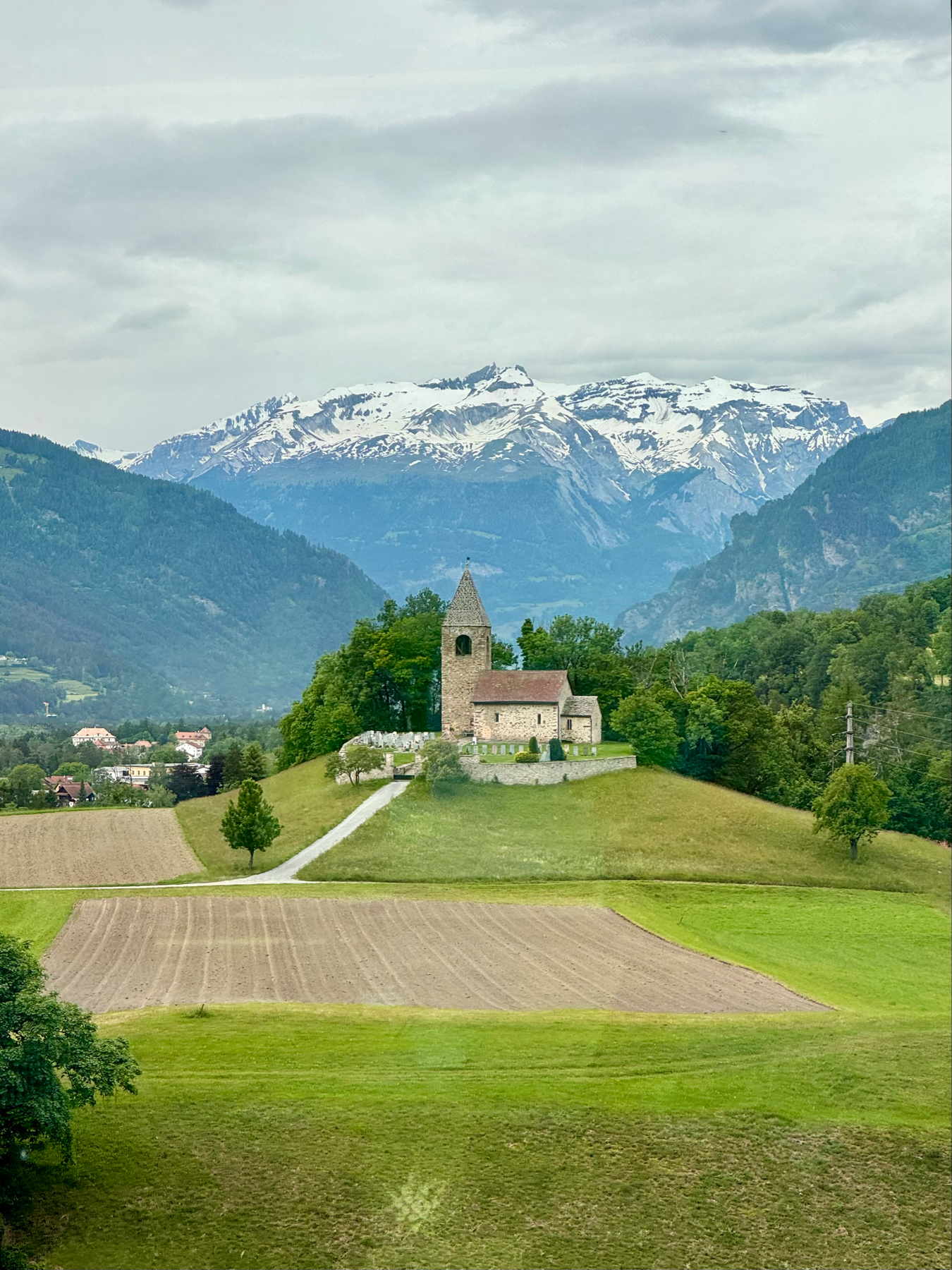 A serene landscape with a small church on a hill, plowed fields in the foreground, and snow-capped mountains in the background.