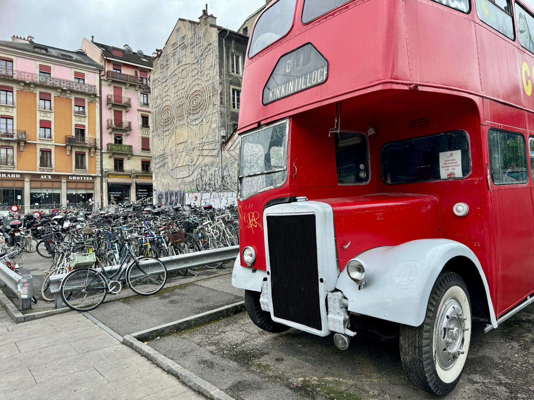 Red double-decker bus parked beside a crowded bicycle rack, with apartment buildings in the background and street art on a building wall.