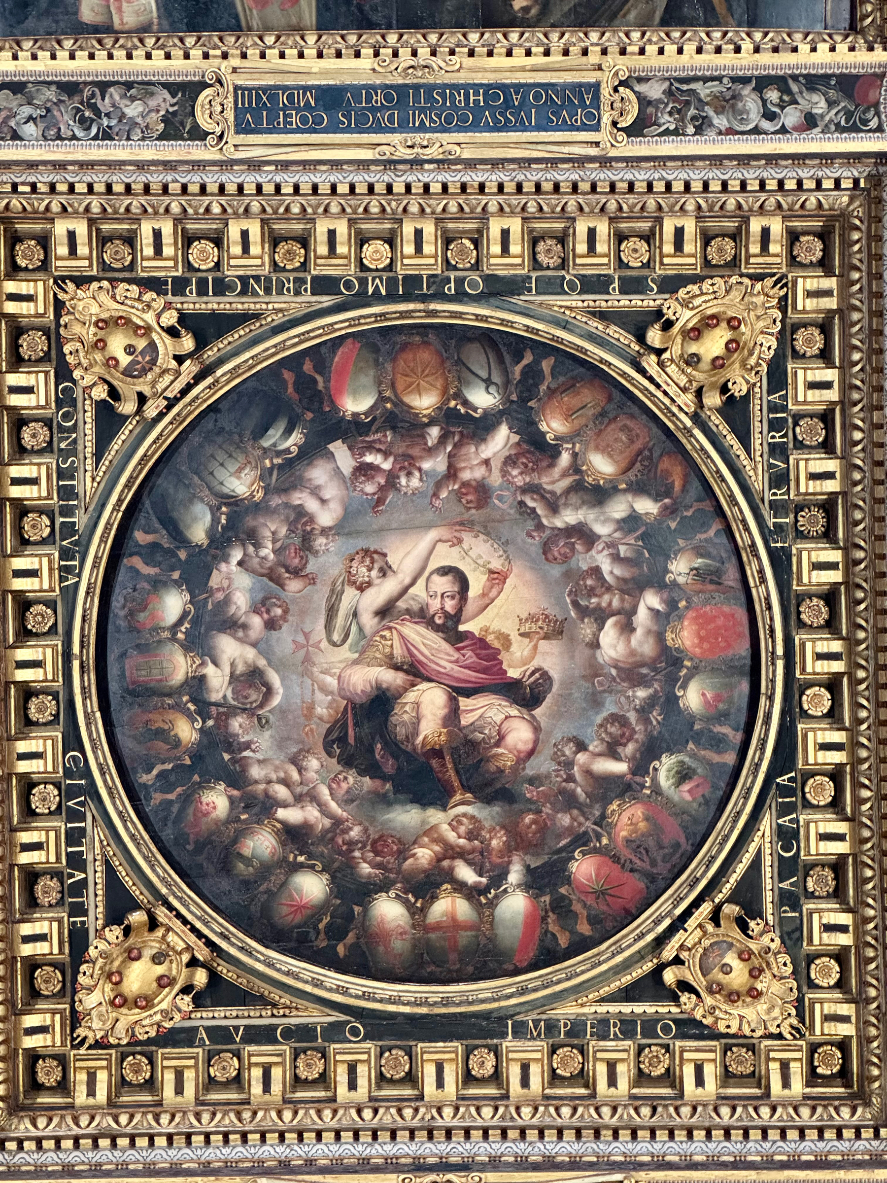 Ornate ceiling painting framed with intricate gold detailing. The central circular painting depicts a regal figure holding a crown, surrounded by cherubs and shields. Latin inscriptions adorn the frame.