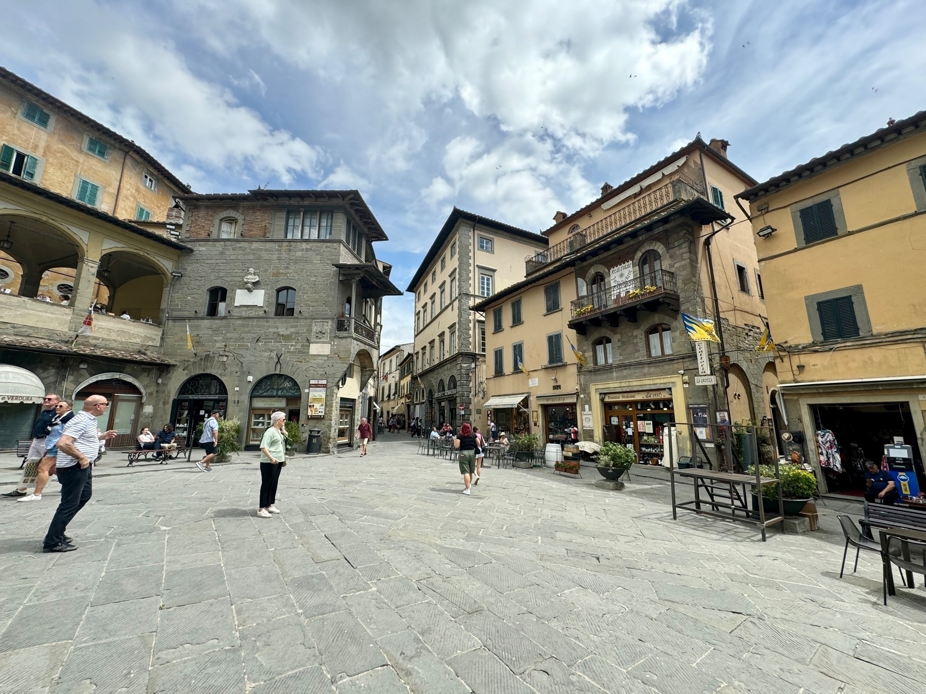 A charming town square with historic buildings and rustic architecture. People stroll and sit at outdoor tables, enjoying the atmosphere. The square is paved with stone tiles, and the buildings feature arched doorways, balconies, and shops at ground level.