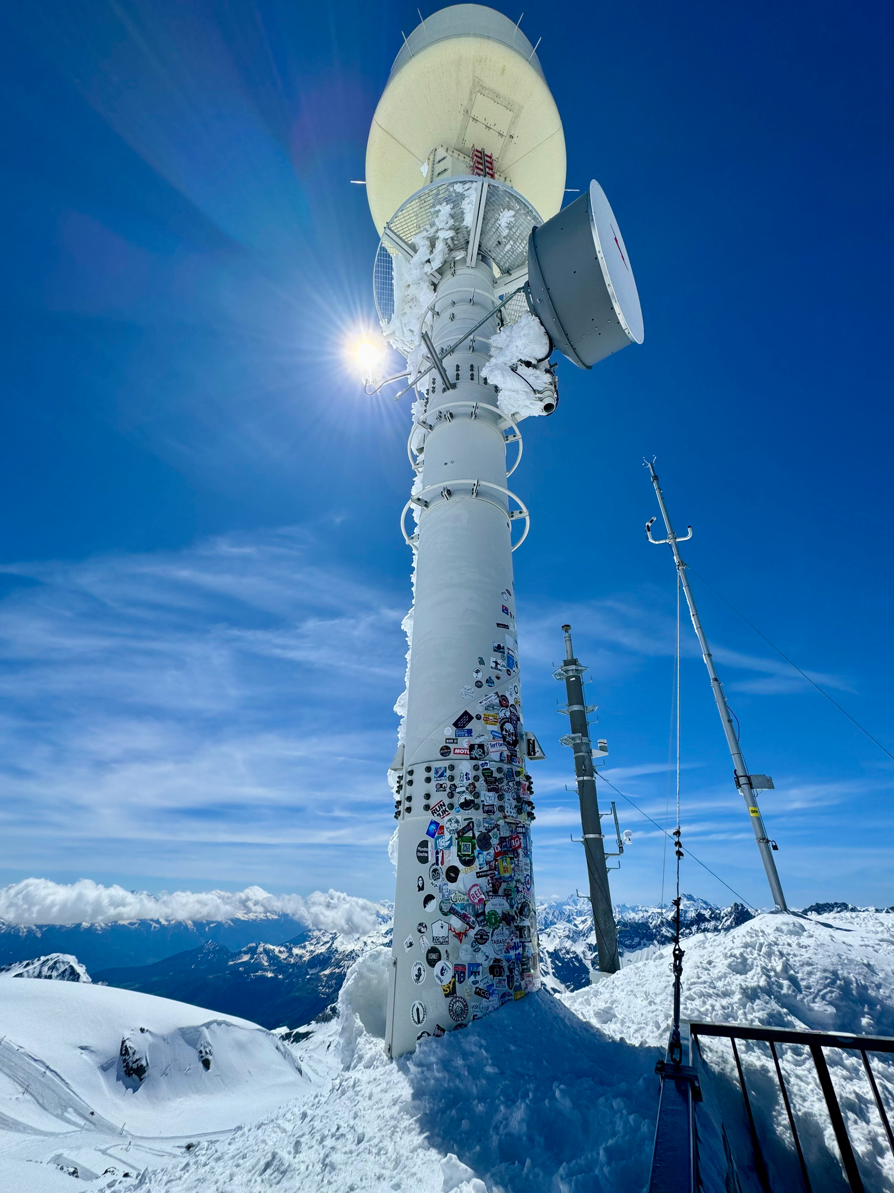 A snow-covered communication tower with numerous stickers on it, set against a clear blue sky with the sun peeking out, surrounded by a snowy mountainous landscape.