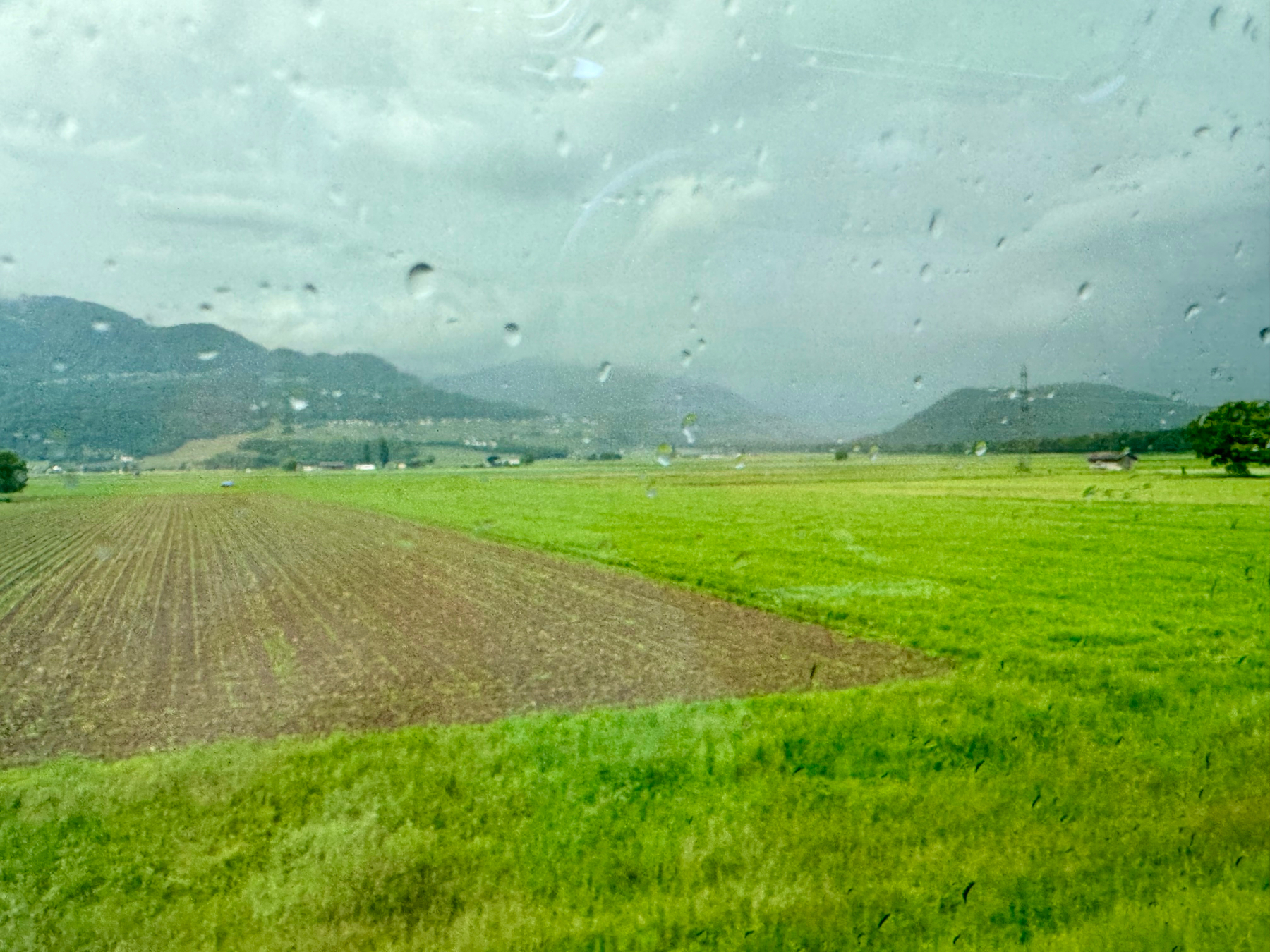 A scenic view of agricultural fields under a cloudy sky, with raindrops visible on the surface from which the photo is taken, suggesting it was shot through a window.