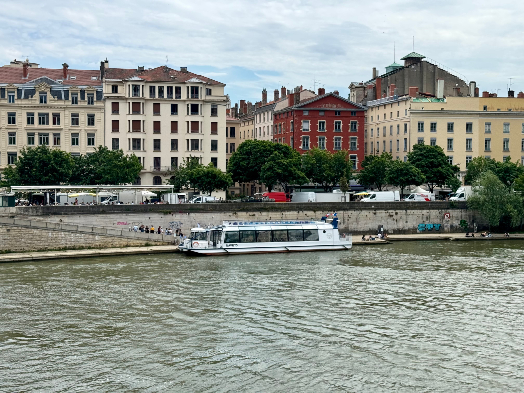 A river with a tour boat, people sitting on the quay, and a backdrop of European-style buildings under a cloudy sky.