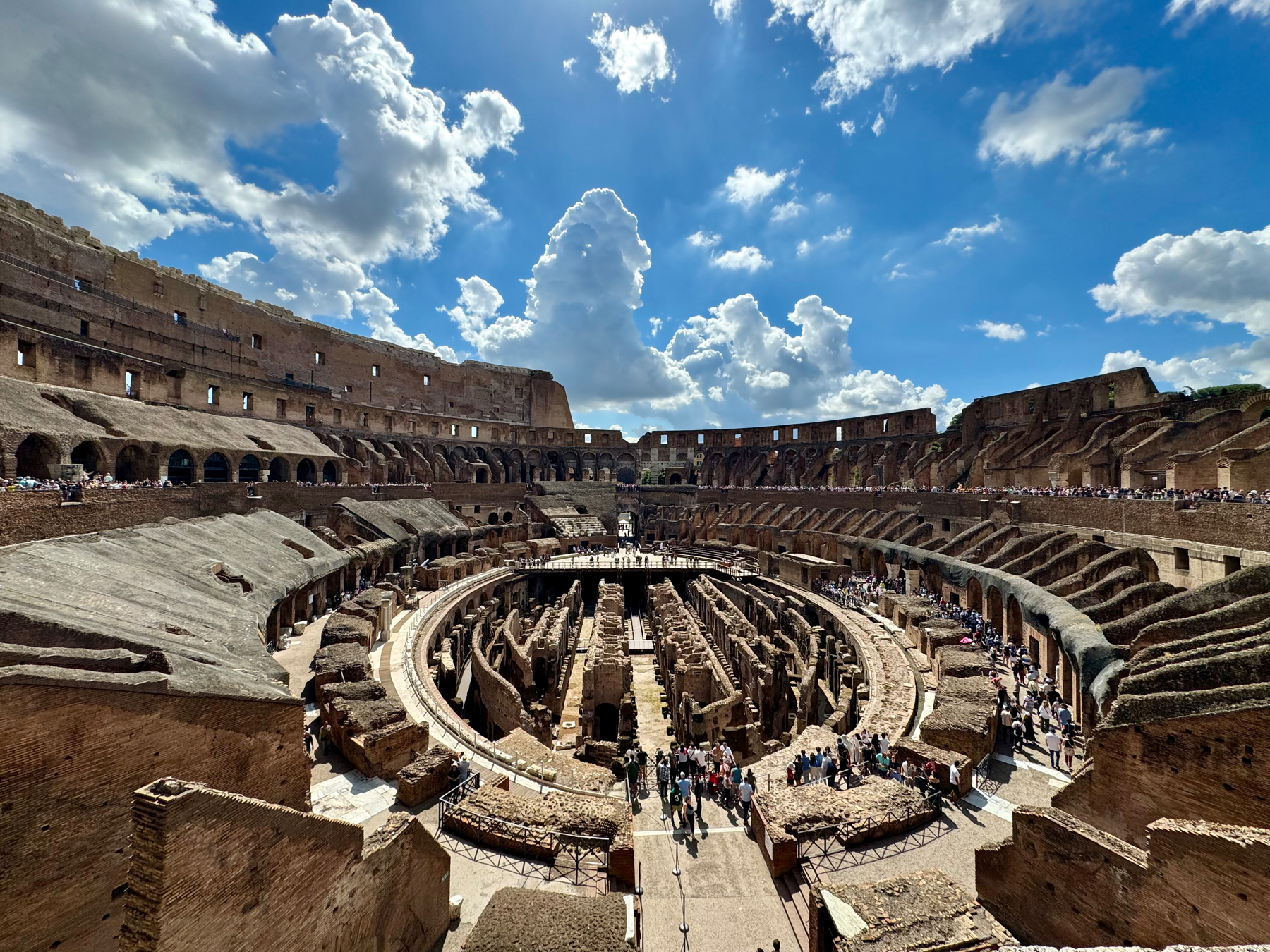 Image of the interior of the Colosseum in Rome, Italy. The structure is partially intact with exposed ancient stone and numerous arches. Visitors are seen walking through the ruins and pathways within the Colosseum under a blue sky filled with scattered clouds.