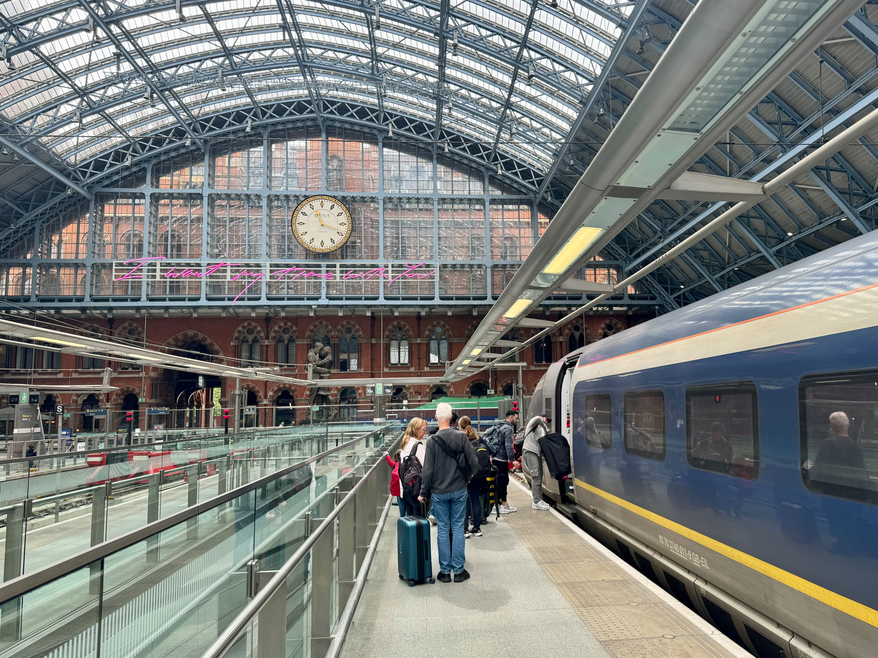 The image shows the St. Pancras train station platform with people boarding a blue train. The station has a large, intricate arched roof made of steel and glass. A clock is prominently displayed above an entrance with a neon sign beneath it.