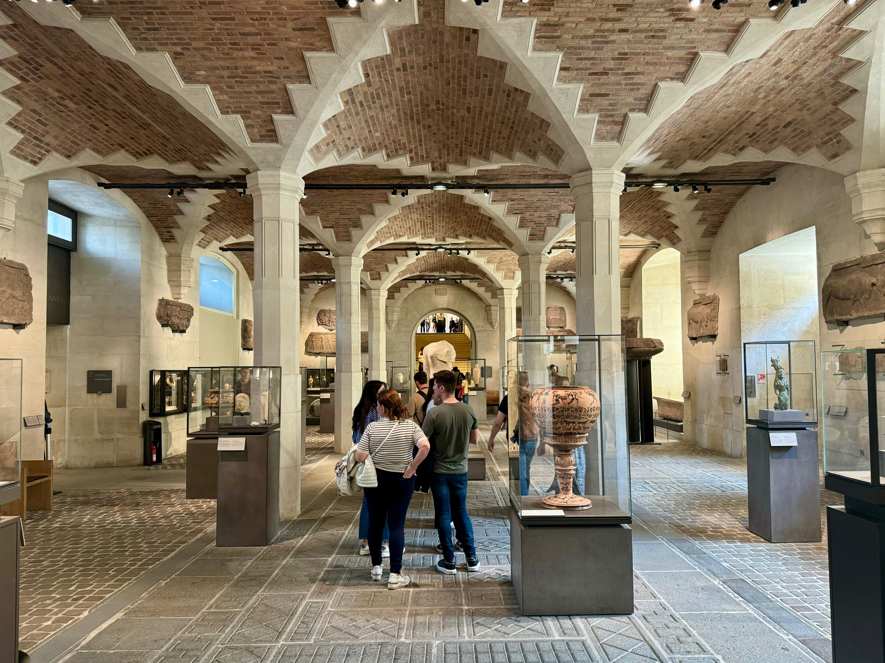 A spacious museum hall with vaulted ceilings, brickwork arches, historical artifacts displayed in glass cases, and visitors observing the exhibits.