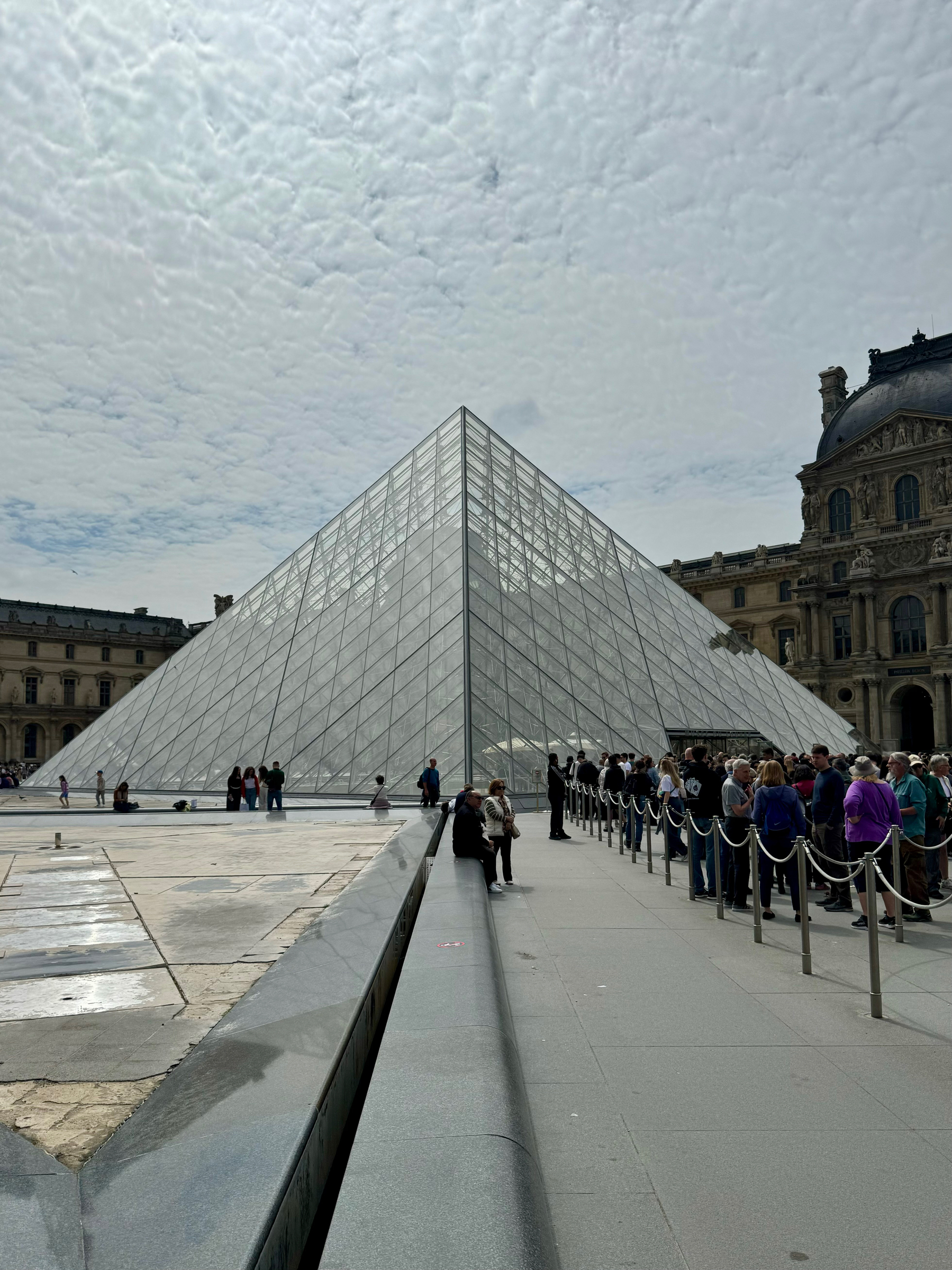 The Louvre Pyramid in Paris on a cloudy day with tourists queuing outside.
