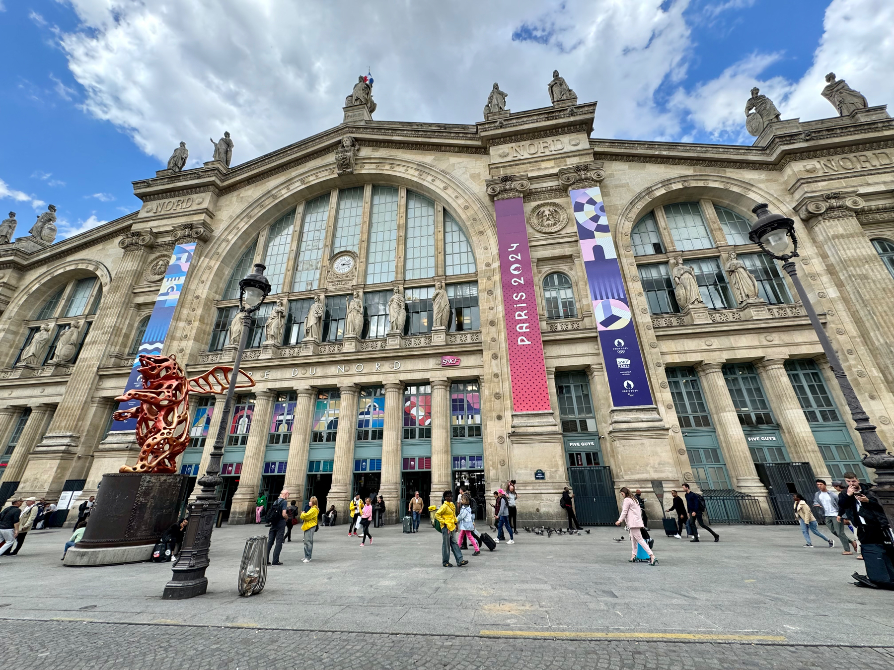 A wide shot of the Gare du Nord train station in Paris with pedestrians walking in front, decorative banners, and a red sculpture near a lamppost.