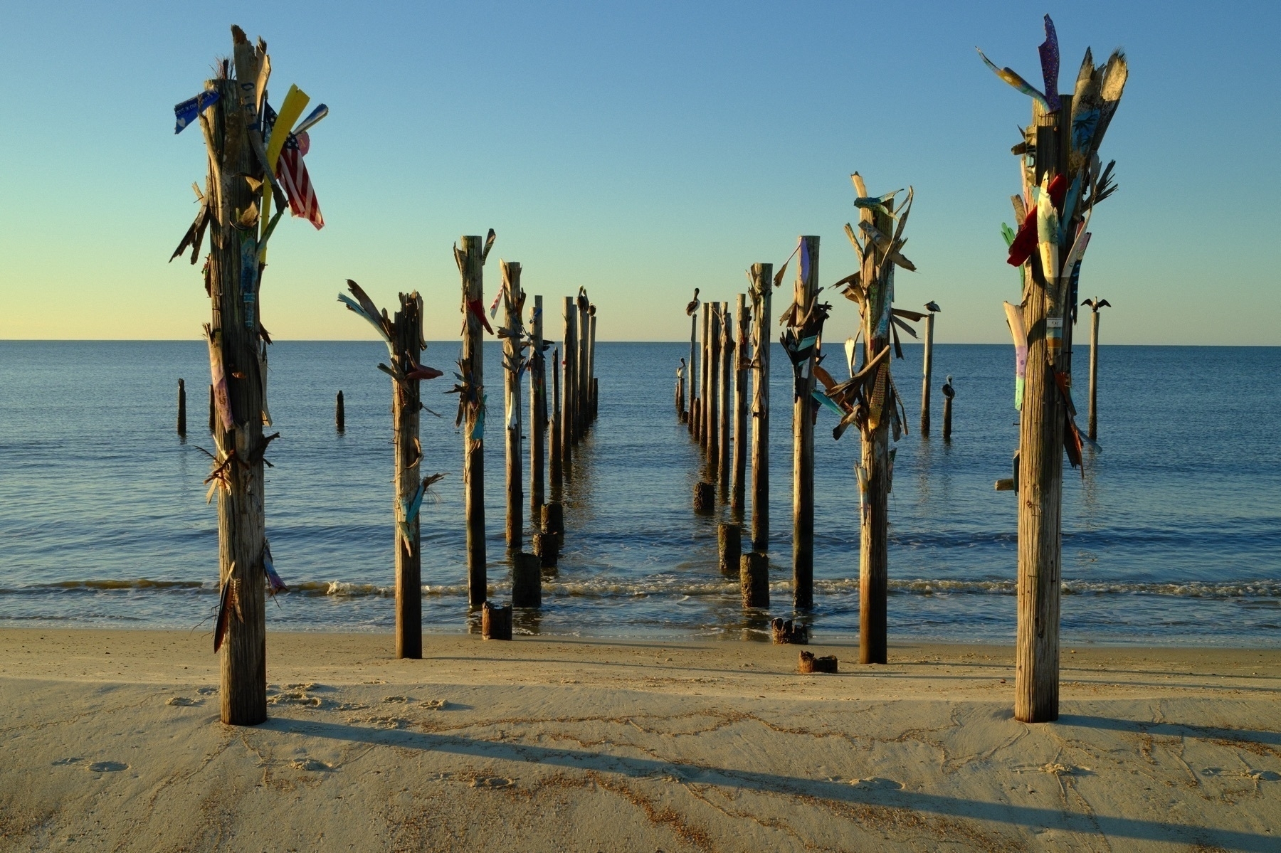 Auto-generated description: Wooden posts adorned with colorful fabric stand in rows leading out into the calm sea from a sandy beach.