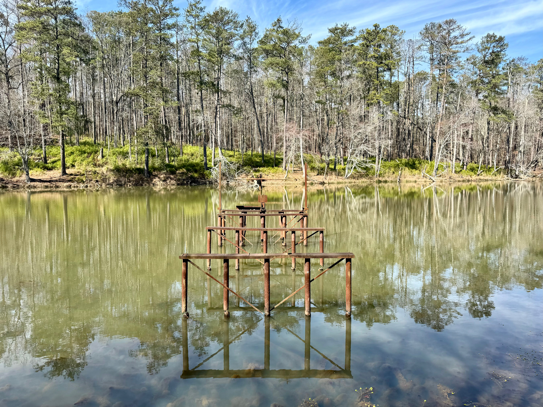 A serene pond with reflections of trees and blue sky, and a rusty metal structure partially submerged in the water.