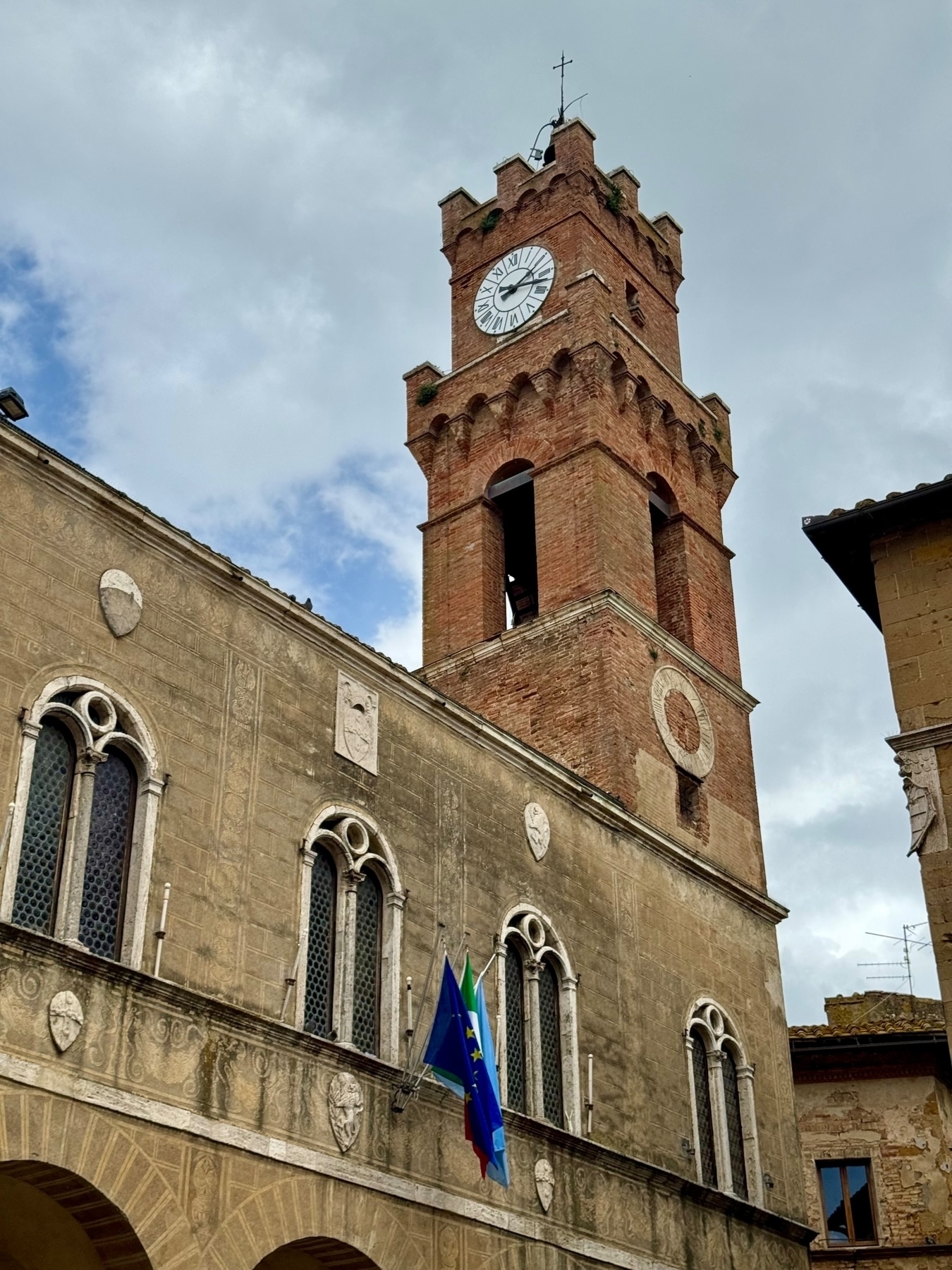 A historic clock tower made of red brick, featuring a white clock face, is attached to an older stone building with arched windows. The building displays Italian and European Union flags. The sky is partly cloudy.